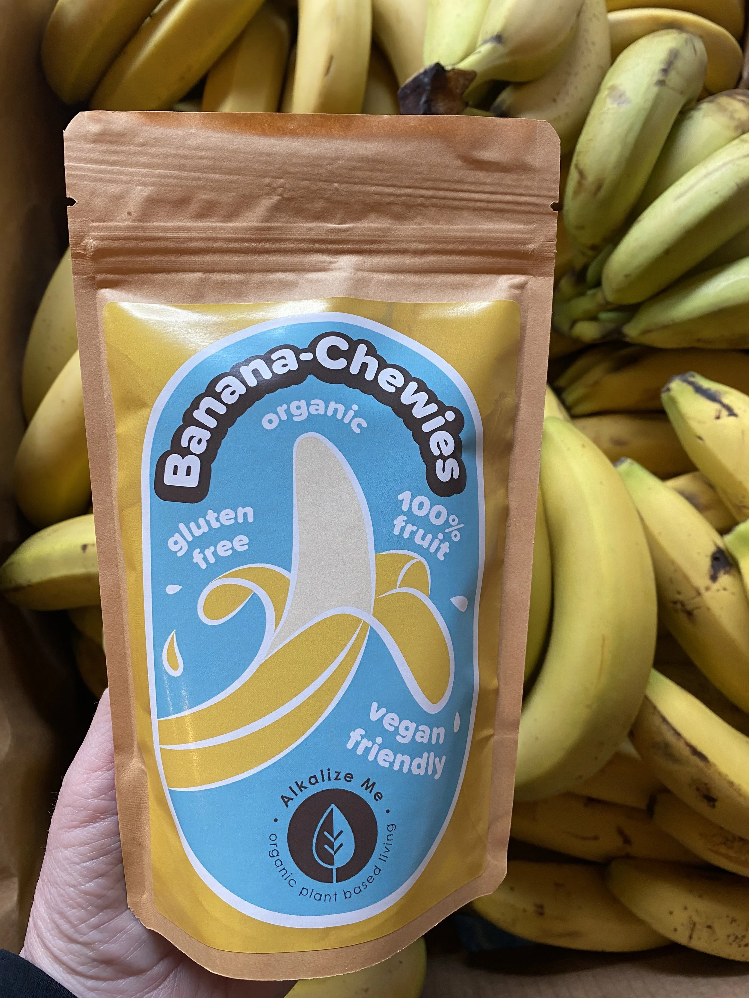 A packet ofAlkalize Me Banana Chewies in front of organic bunches of ripe bananas.
