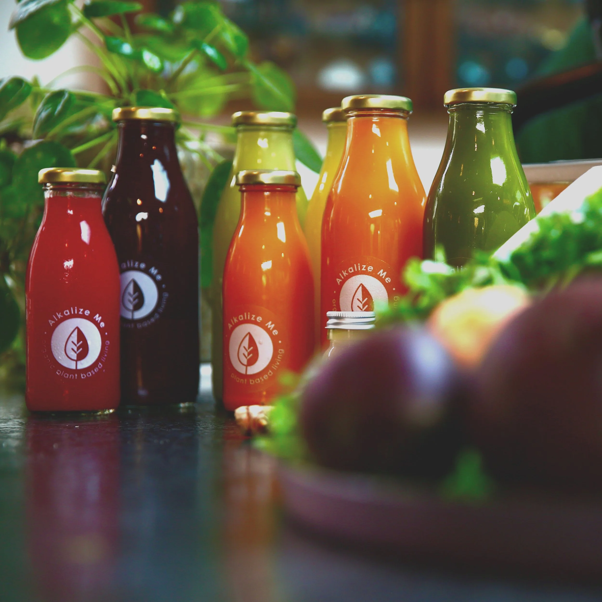 Variety of Alkalize Me cold-pressed juice bottles in a kitchen setting with a bowl of vegetables in the foreground.