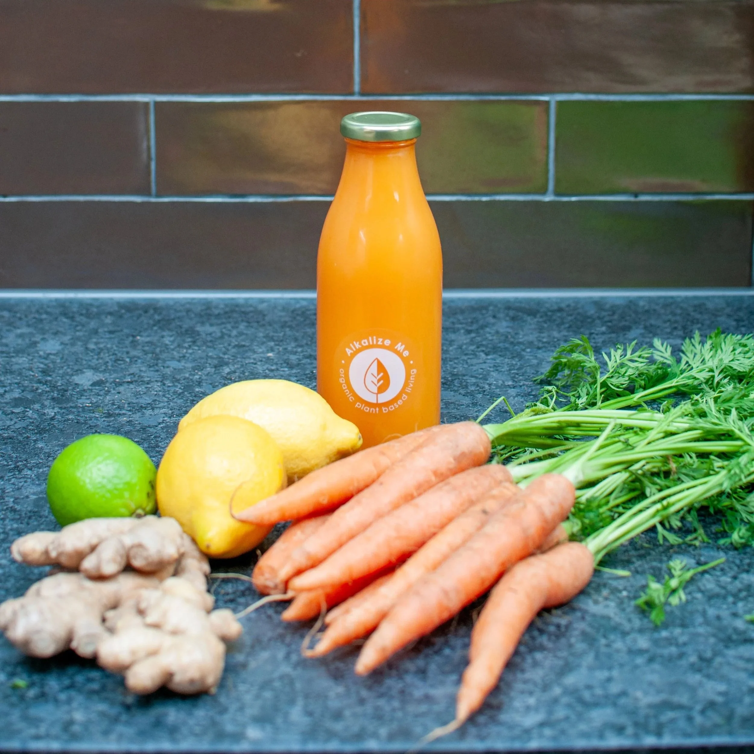 A glass bottle containing Night Vision, a cold-pressed juice made of organic carrots, lemon, lime and ginger. On a kitchen counter surrounded by the fresh, organic ingredients.