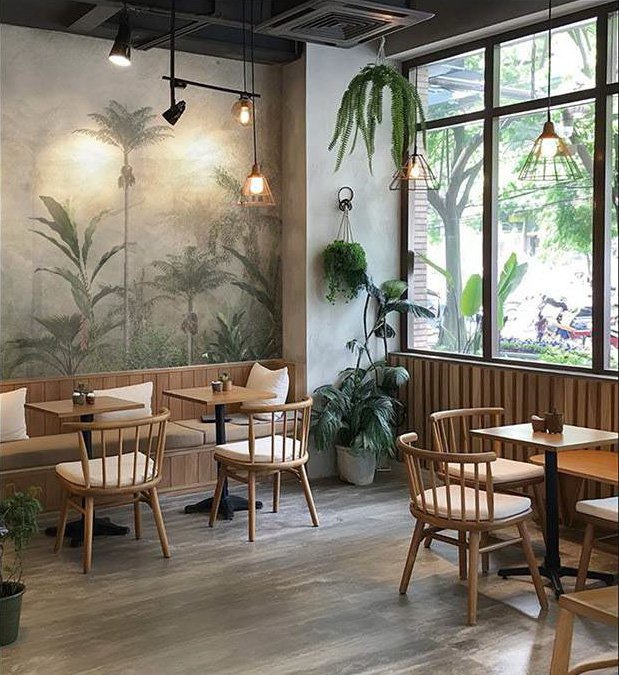 Cozy cafe interior with wooden tables and chairs, wall mural of large plants, hanging pendant lights, and potted plants near large window with a view of trees outside.