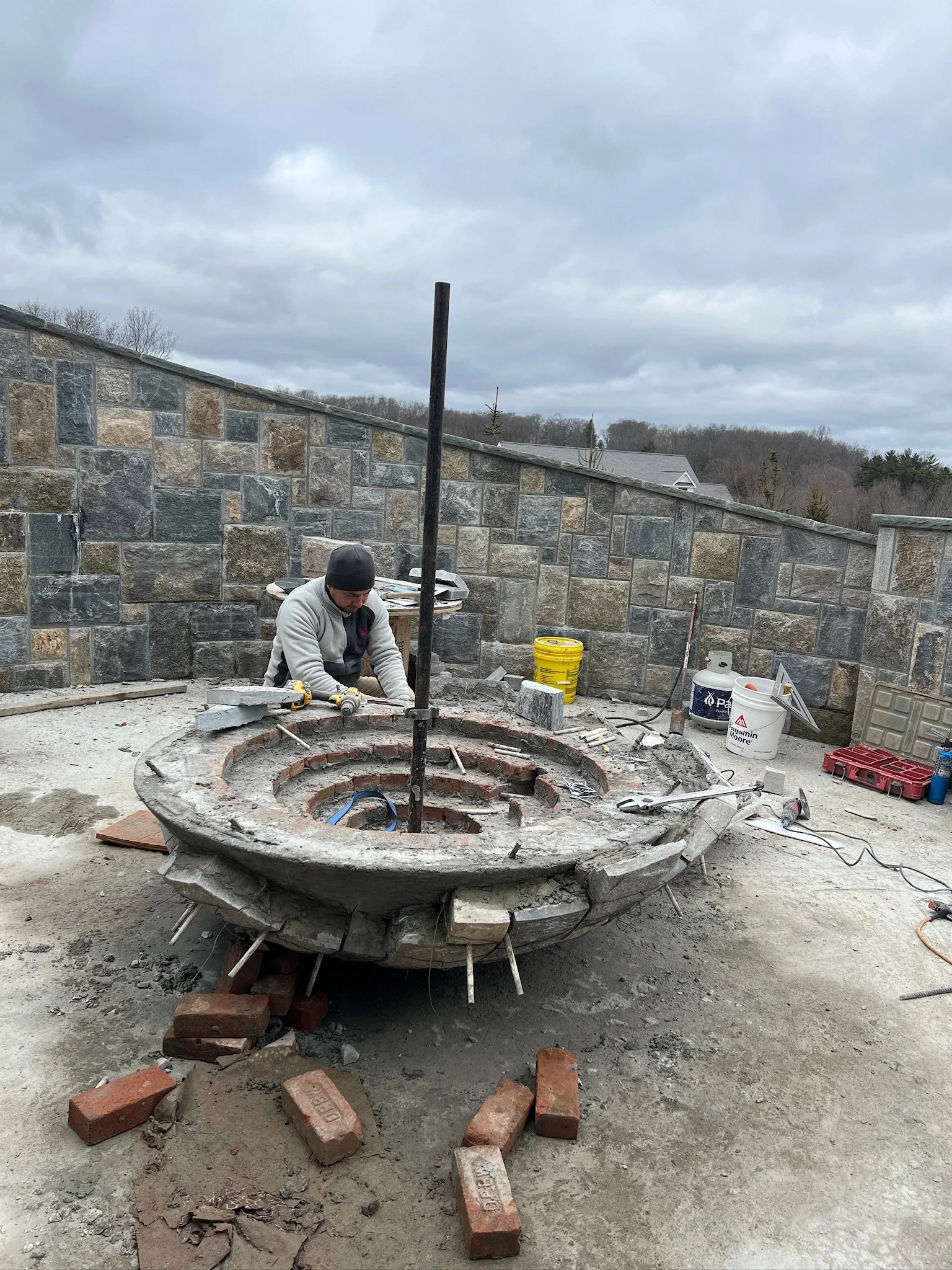 A construction worker working on a circular brick and concrete fire pit in an outdoor patio area, with tools and construction materials around him, surrounded by a stone wall and cloudy sky.