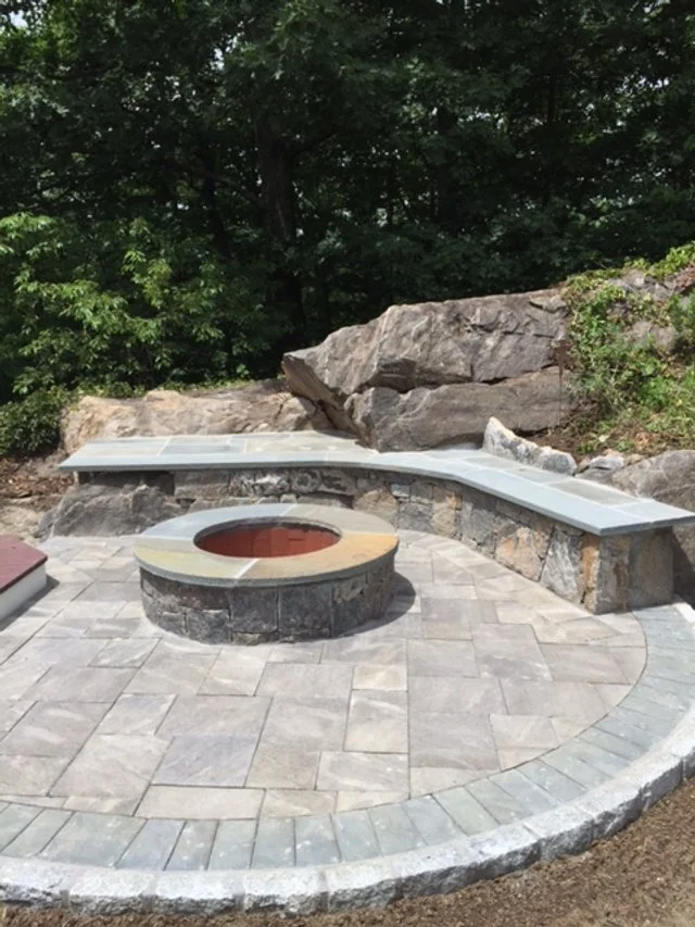 Outdoor fire pit with gray stone surround, set in a paved patio area, with a curved stone bench and large rocks in the background, surrounded by trees.