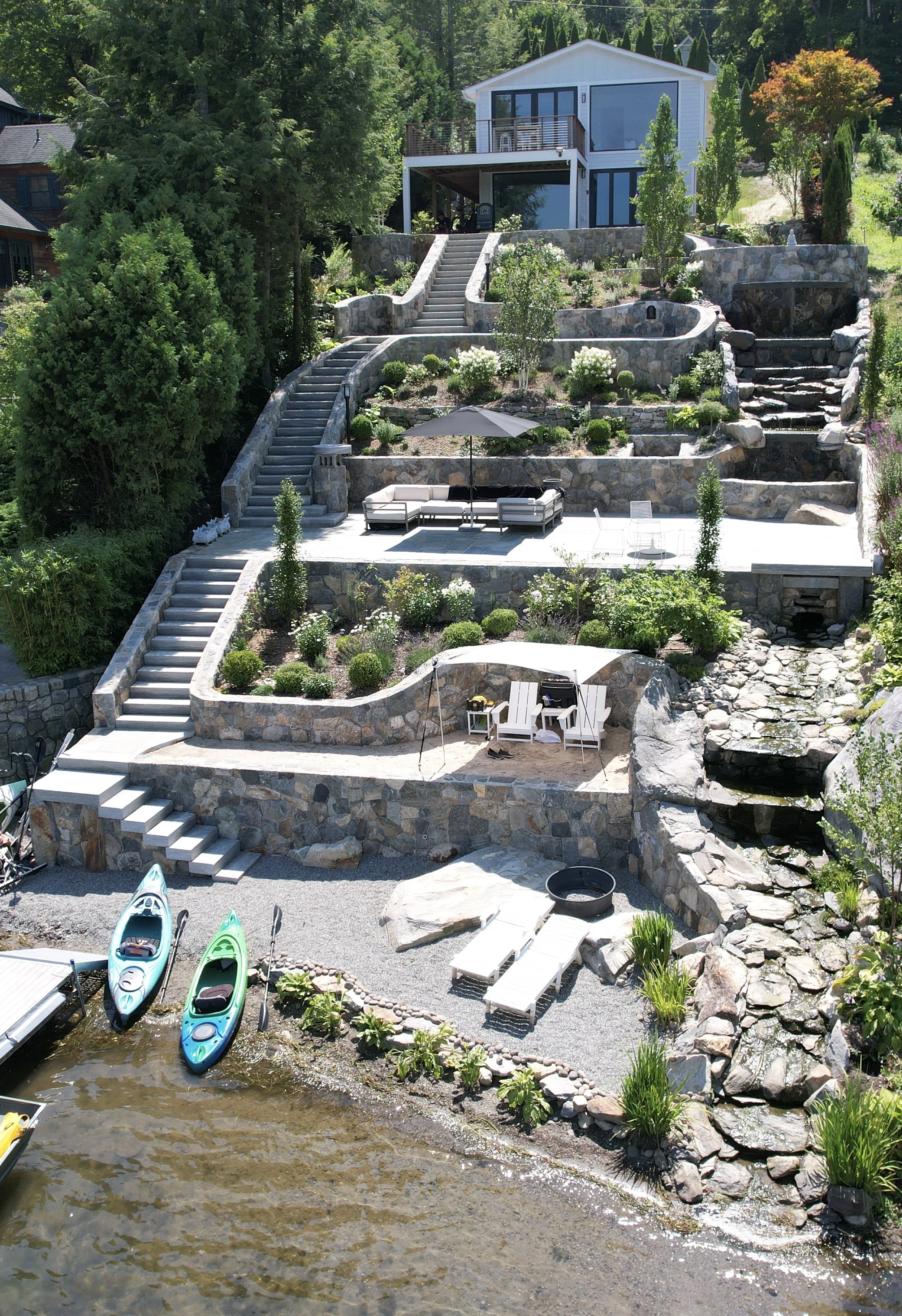 Multi-level waterfront backyard with stone terraces, stairs, lush vegetation, a patio with seating, umbrellas, kayaks near the water, and a modern house at the top of the hill.
