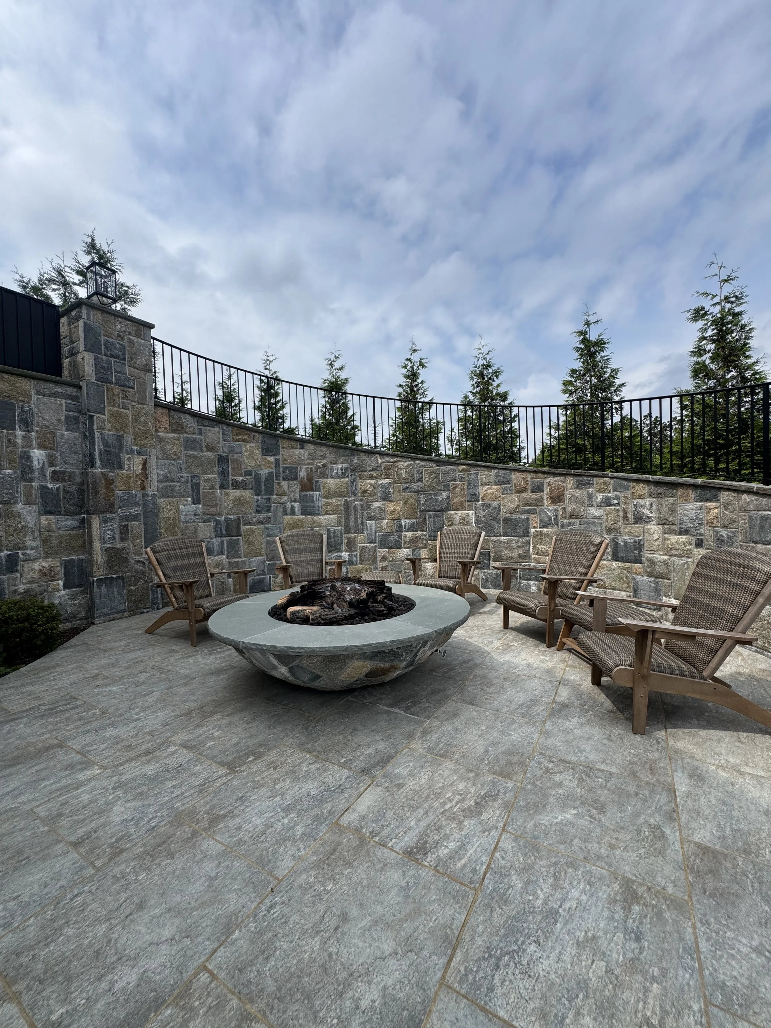 Outdoor patio with a circular fire pit in the center surrounded by six wooden and wicker chairs, enclosed by a stone wall and black metal railing, with a cloudy sky and trees in the background.