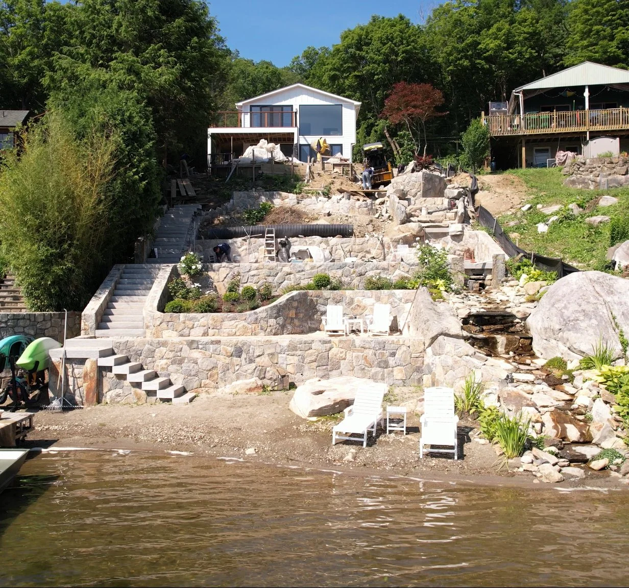 Construction work on a lakeside residential property, with stairs, rocks, and water in the foreground, and a house and trees in the background.