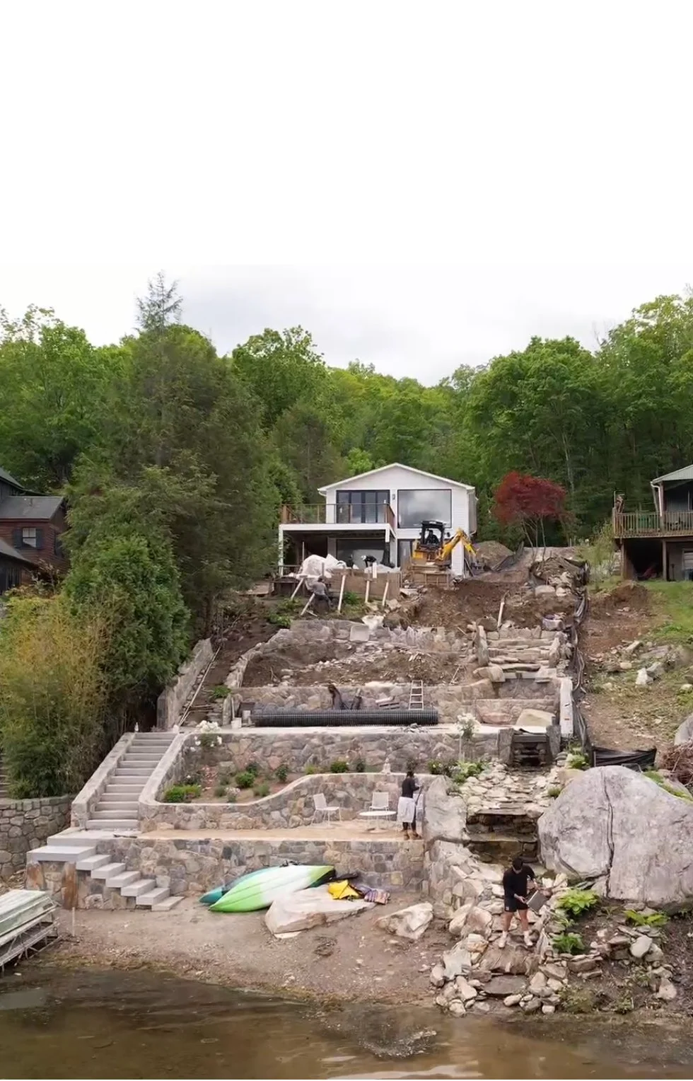Construction site on a hillside next to a body of water. Stone stairs and retaining walls are being built, with workers present. There is a small white house at the top of the hill and construction equipment around.