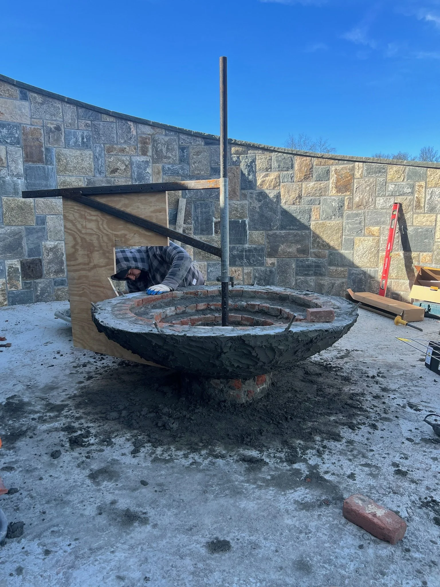 Construction worker installing a brick-based fire pit on a concrete patio outside, with tools and building materials nearby and a stone wall in the background.