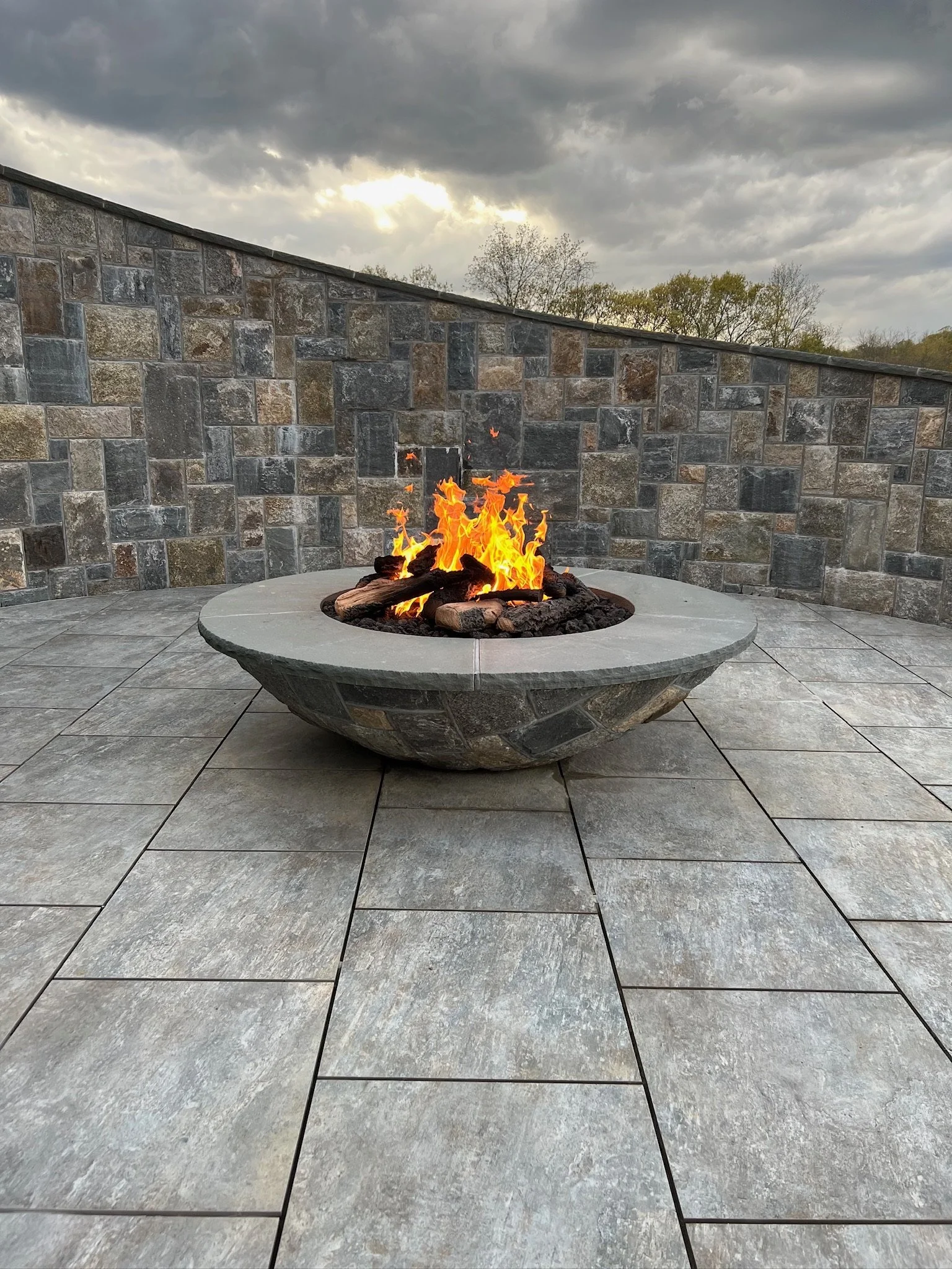 A circular stone fire pit with burning logs on a patio, surrounded by a stone wall, with overcast sky overhead.