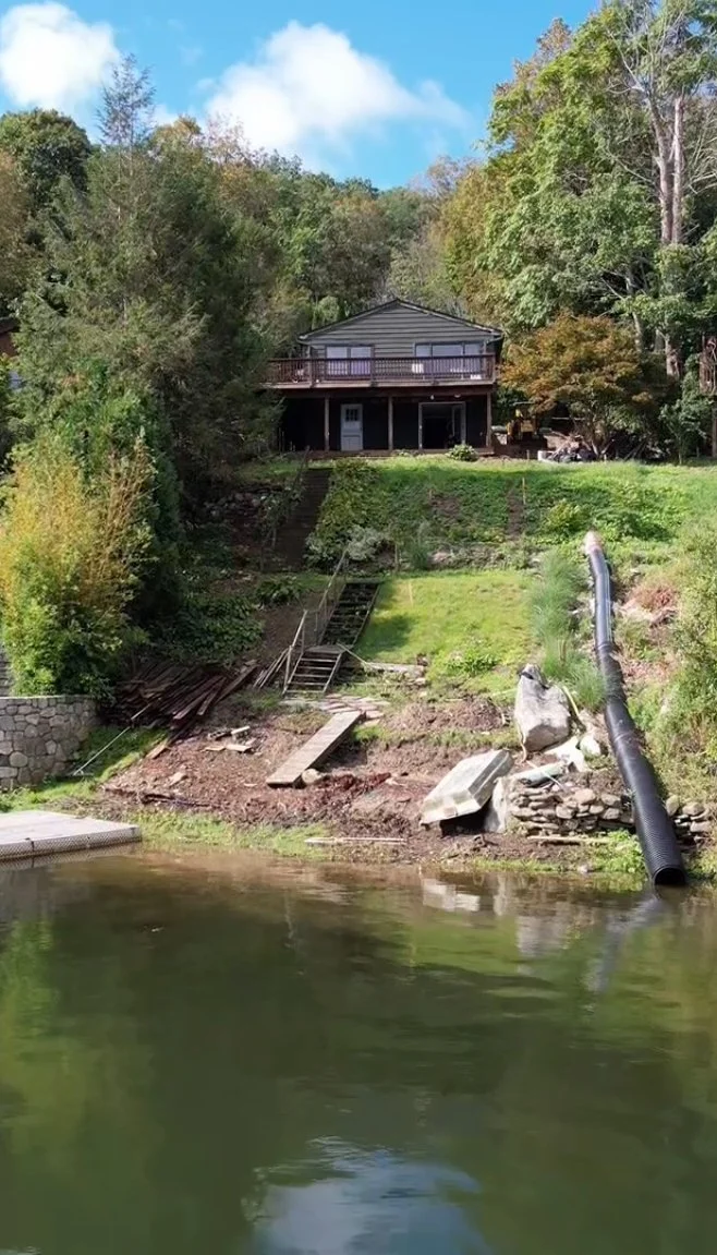A house on a hill overlooking a body of water, with construction materials and pipes visible near the shore, and trees surrounding the area.