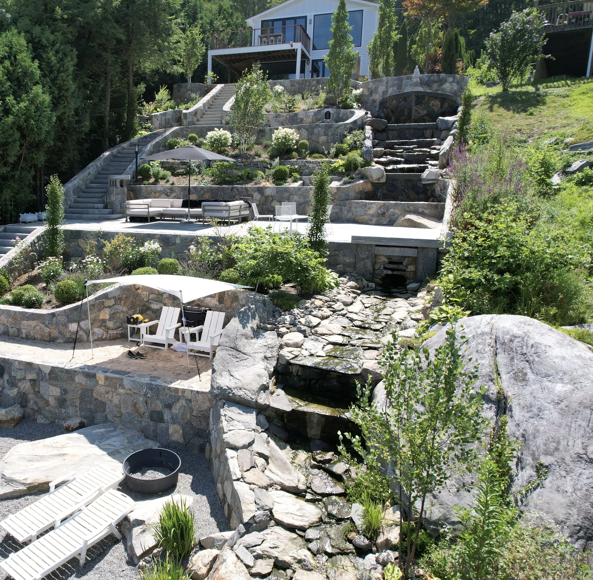 Multi-level landscaped backyard with stone terraces, stairs, waterfalls, a stream, and secluded seating areas with umbrellas and lounge chairs.