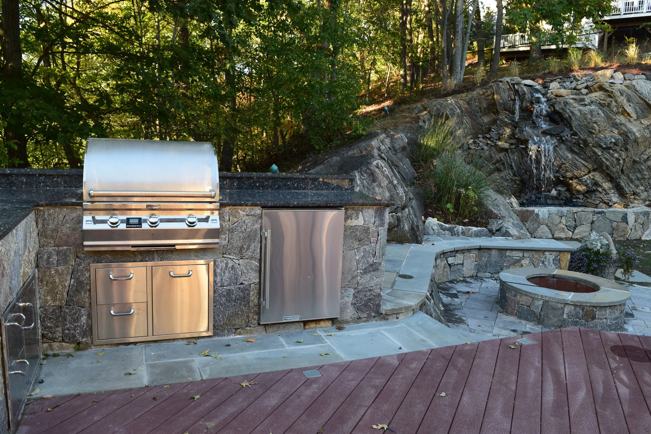 Outdoor kitchen with stainless steel grill, small fridge, and storage cabinets built into a stone countertop, with a landscaped backdrop including a water feature and trees.