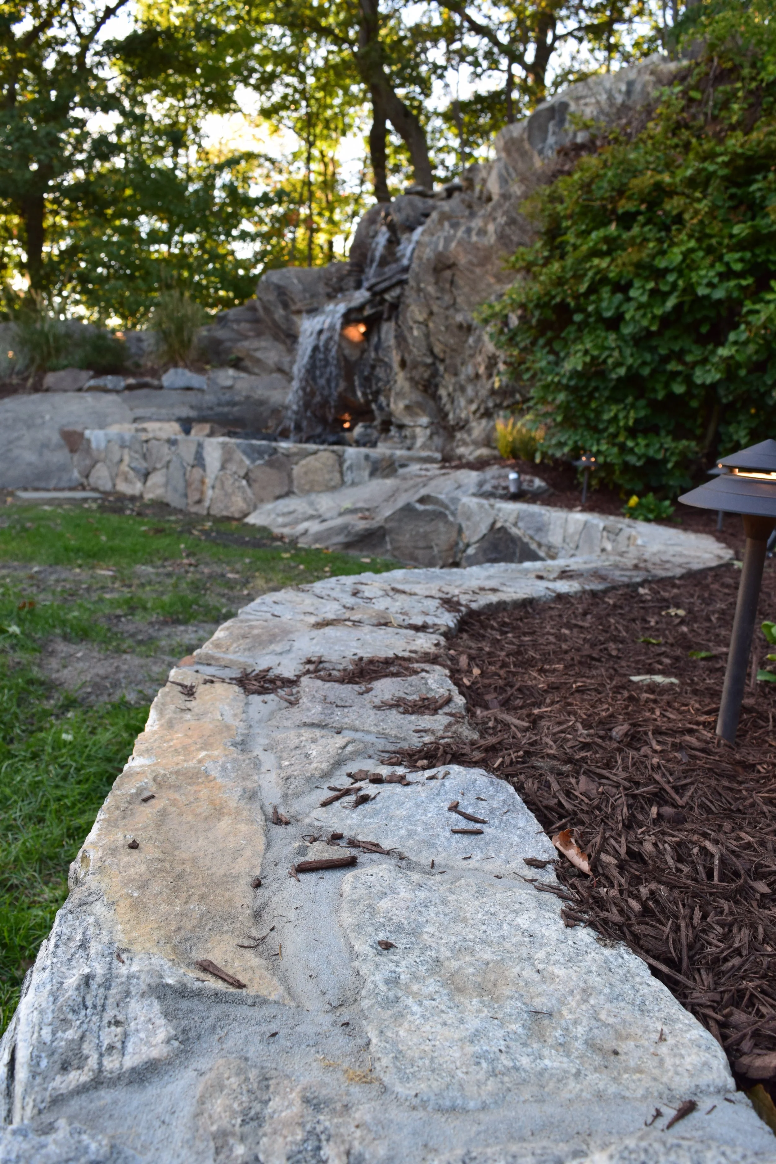 Stone pathway and garden with a small waterfall feature surrounded by trees and landscape lighting.