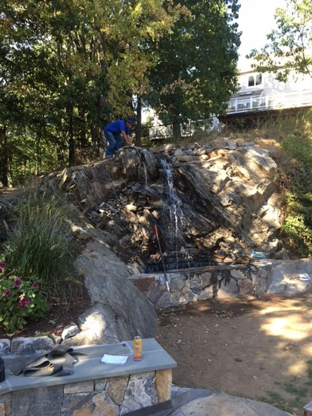 A person working on a small water feature or waterfall built into a rock hillside in a backyard.