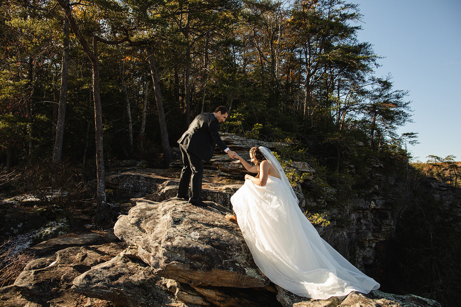 Groom stands on a rock formation while he helps his bride step up to join him during their elopement by Promise Mountain