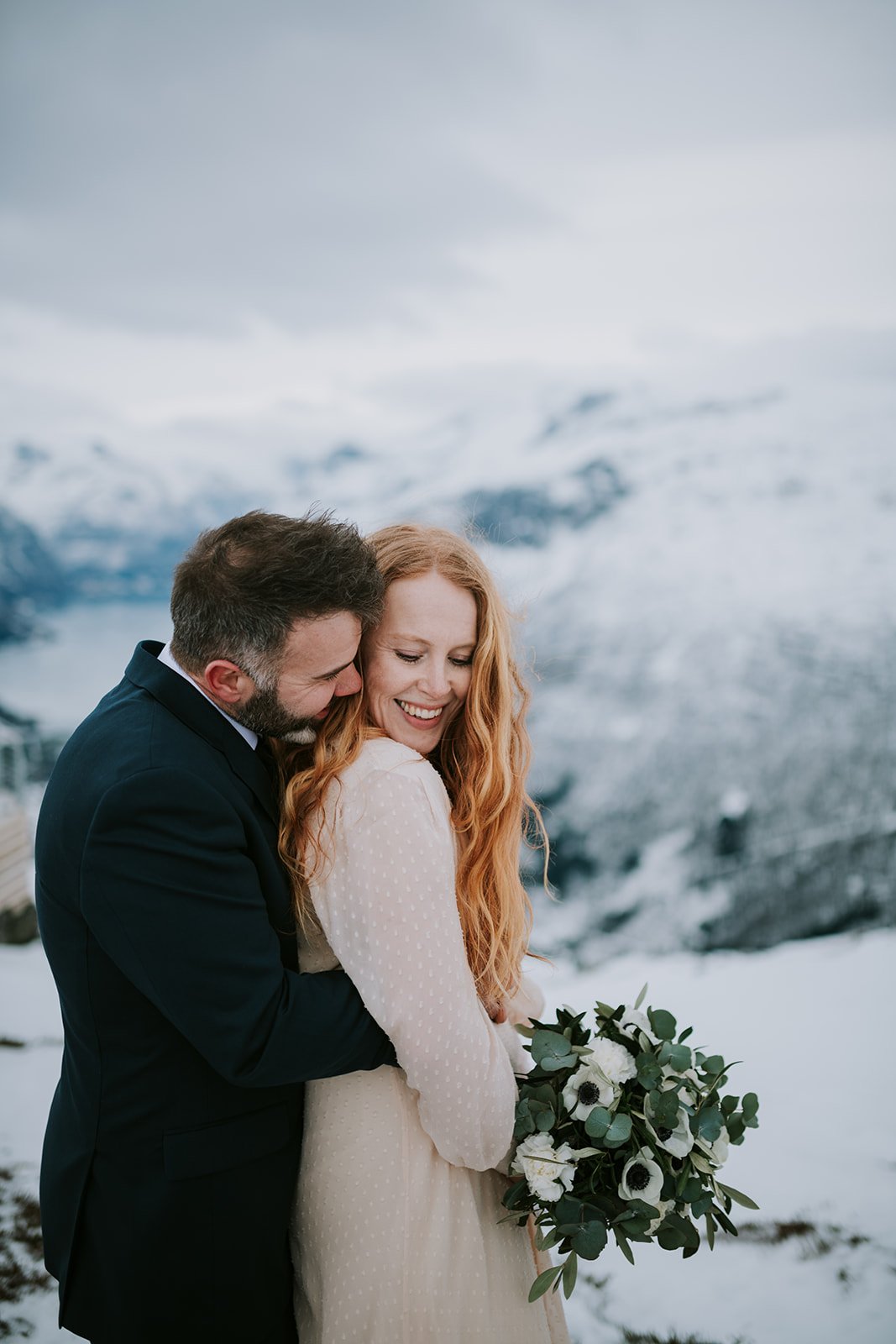 Groom hugs the bride from behind as she smiles while holding her bouquet at a snowy elopement destination