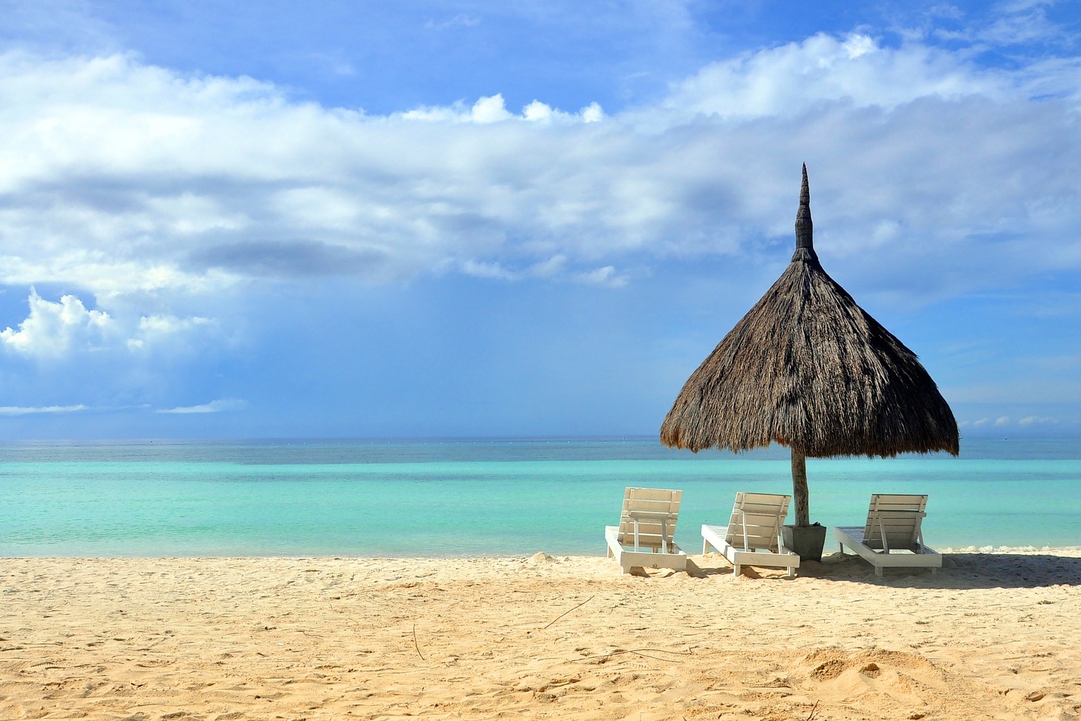 A grass hut and lounge chairs sitting on a blissfully empty beach in front of the brilliant blue water.