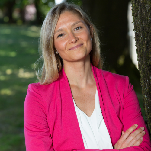 Woman with blonde hair wearing a pink blazer and white top, standing outdoors by a tree with a park in the background.