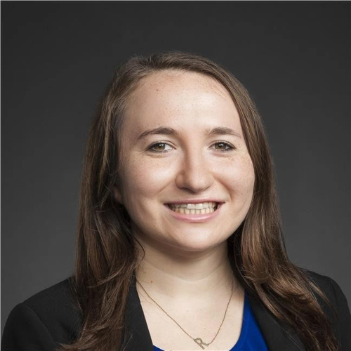 Headshot of a young woman with long brown hair, smiling, wearing a black blazer and blue top against a dark gray background.