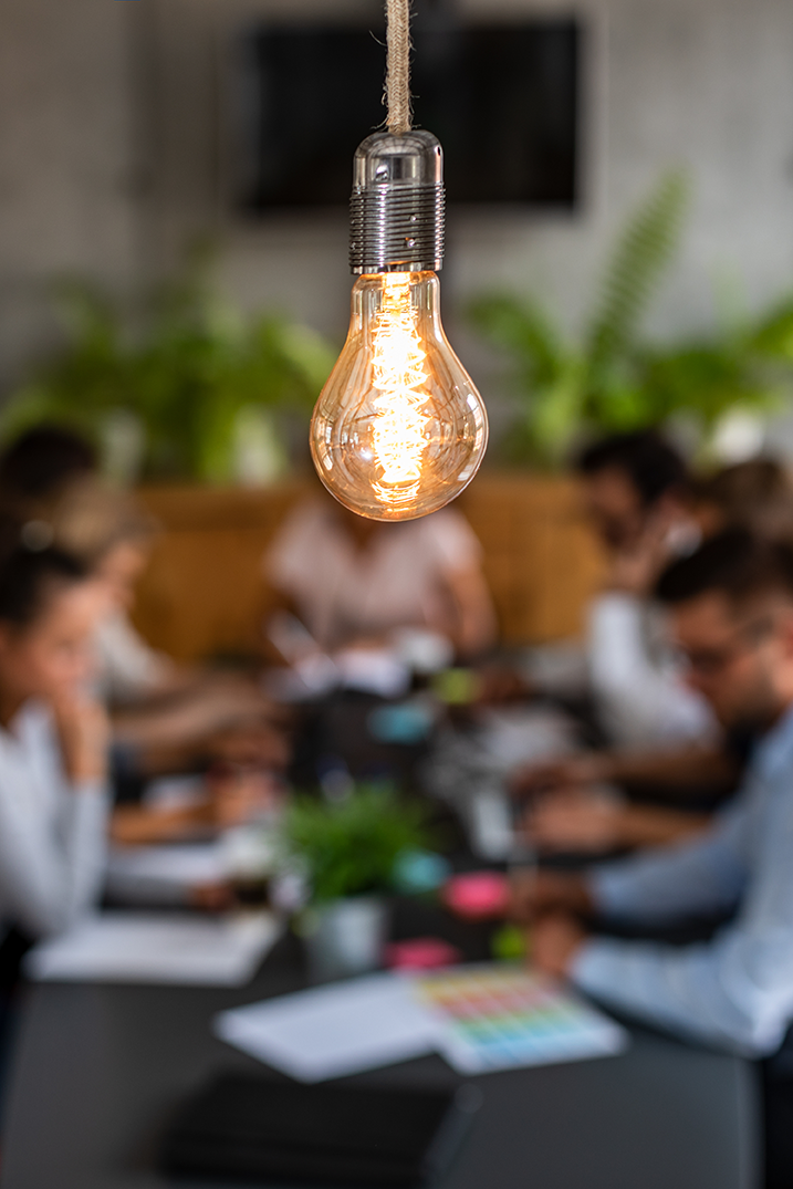 Close-up of an illuminated vintage-style light bulb hanging from a string in a meeting room with blurred people working at a table and a TV or monitor on the wall in the background.