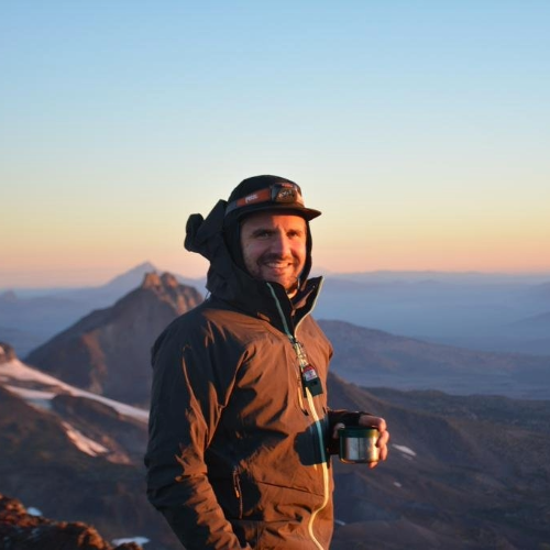 Nick Nilo, NMCG's Senior Regulatory Affairs Specialist, standing on mountain at sunset with view of distant mountains and snow patches.