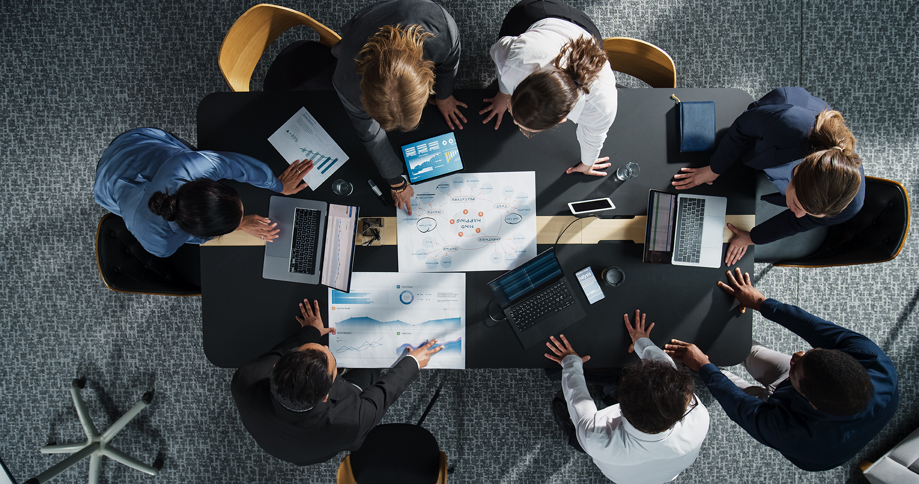 Top view of a business meeting with eight professionals gathered around a conference table discussing charts and graphs displayed on laptops, tablets, and paper documents.