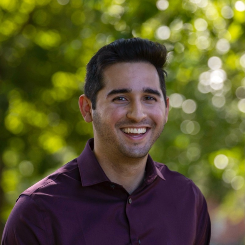 A smiling man with dark hair wearing a dark purple shirt outdoors with green trees in the background.