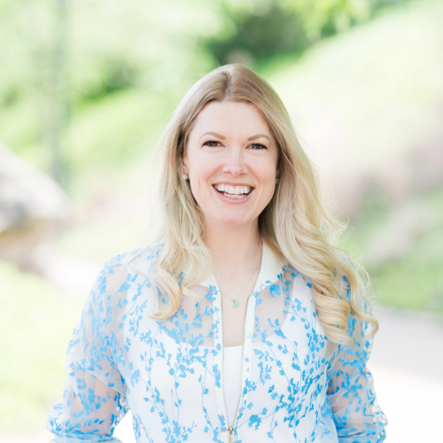 A smiling woman outdoors with blonde hair, wearing a light blue and white patterned jacket and a white top.