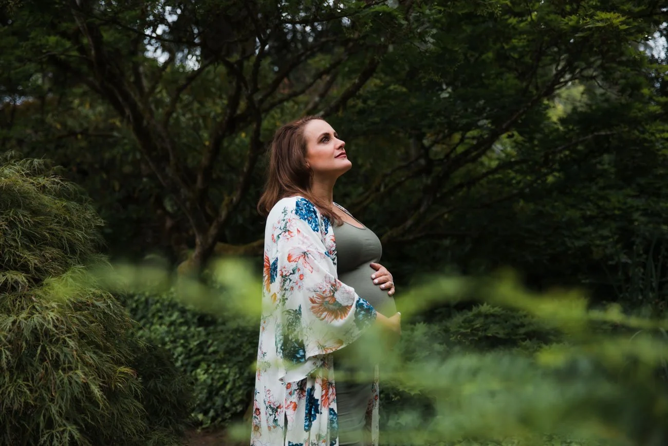 A pregnant woman standing outdoors in a lush, green garden, looking up thoughtfully, with her hand resting on her belly.