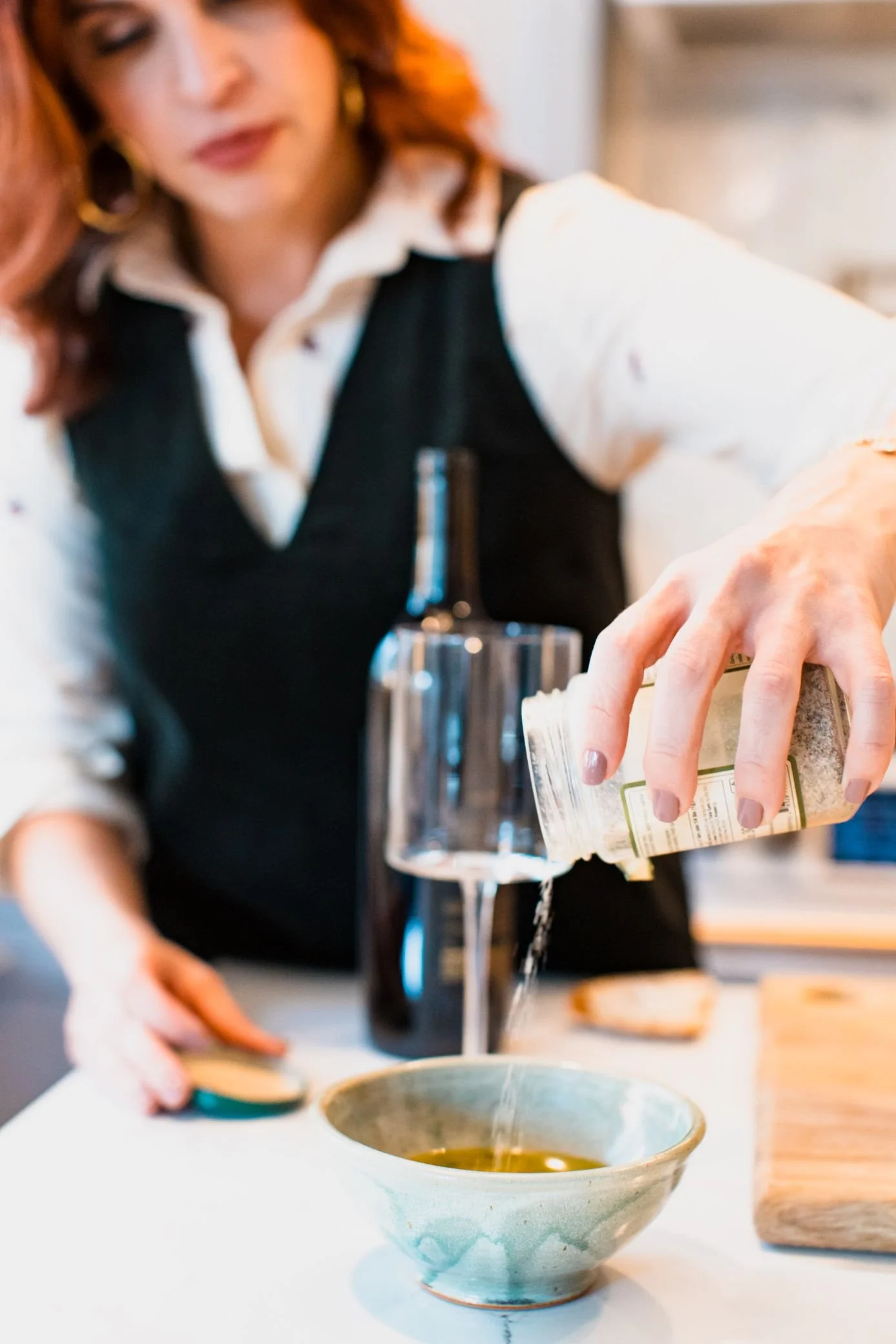 Woman pouring oil into a bowl in a kitchen, with a bottle and wooden cutting board nearby.