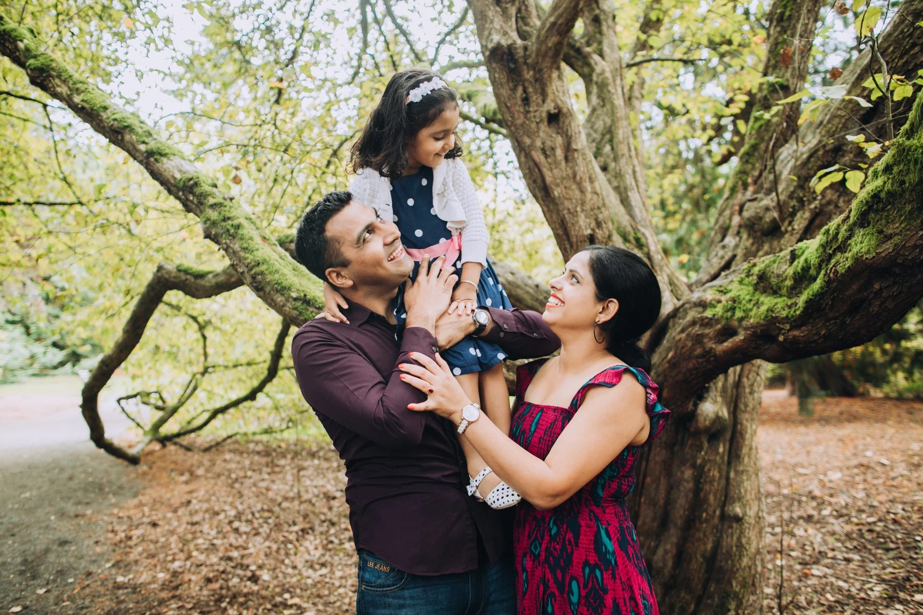 A family of three, consisting of a man, woman, and young girl, standing outdoors in front of a large tree with green leaves, smiling and enjoying their time together.
