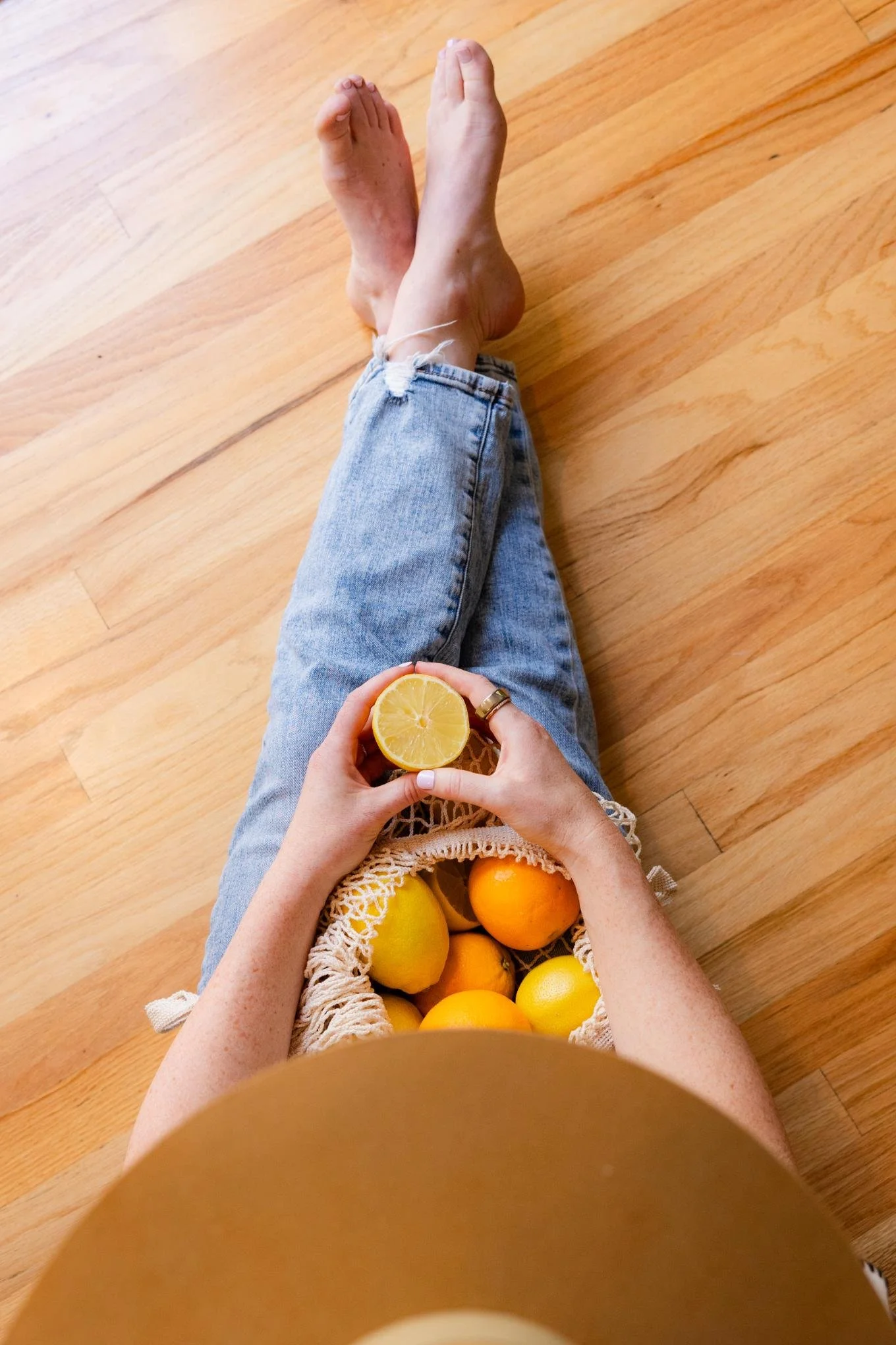 Person sitting on the floor with legs extended, holding a lemon half in hand, with a basket of lemons and oranges on their lap, on a wooden floor.