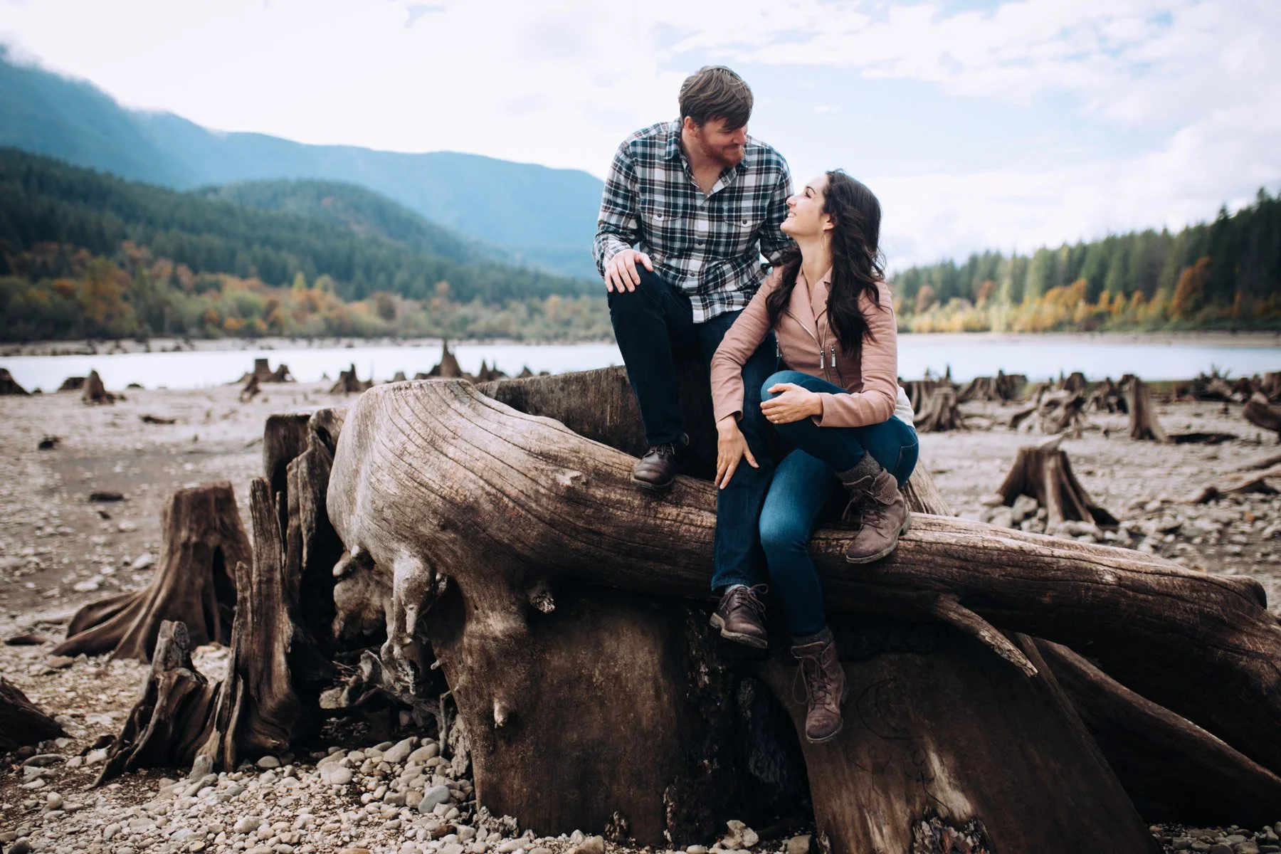 A man and woman sitting on a large fallen tree trunk on a rocky lakeshore, with mountains and trees in the background, smiling and looking at each other.