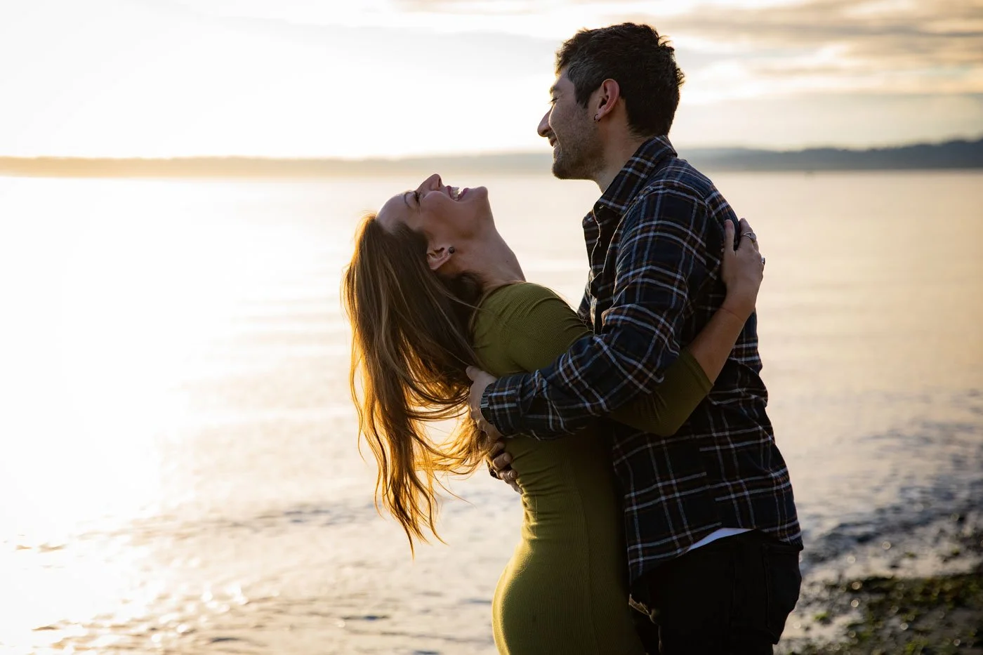 A couple at the beach during sunset, with the woman in an olive-green dress and the man in a plaid shirt, embracing and smiling at each other.