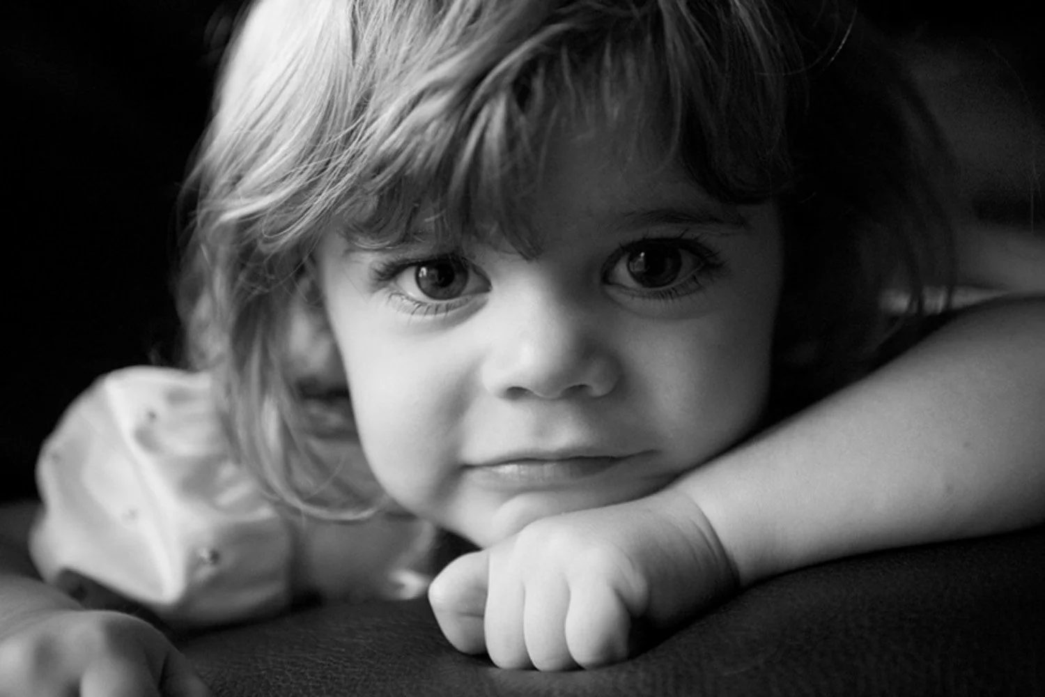 Black and white close-up photo of a young girl with curly hair resting her chin on her arm, looking at the camera with a calm expression.