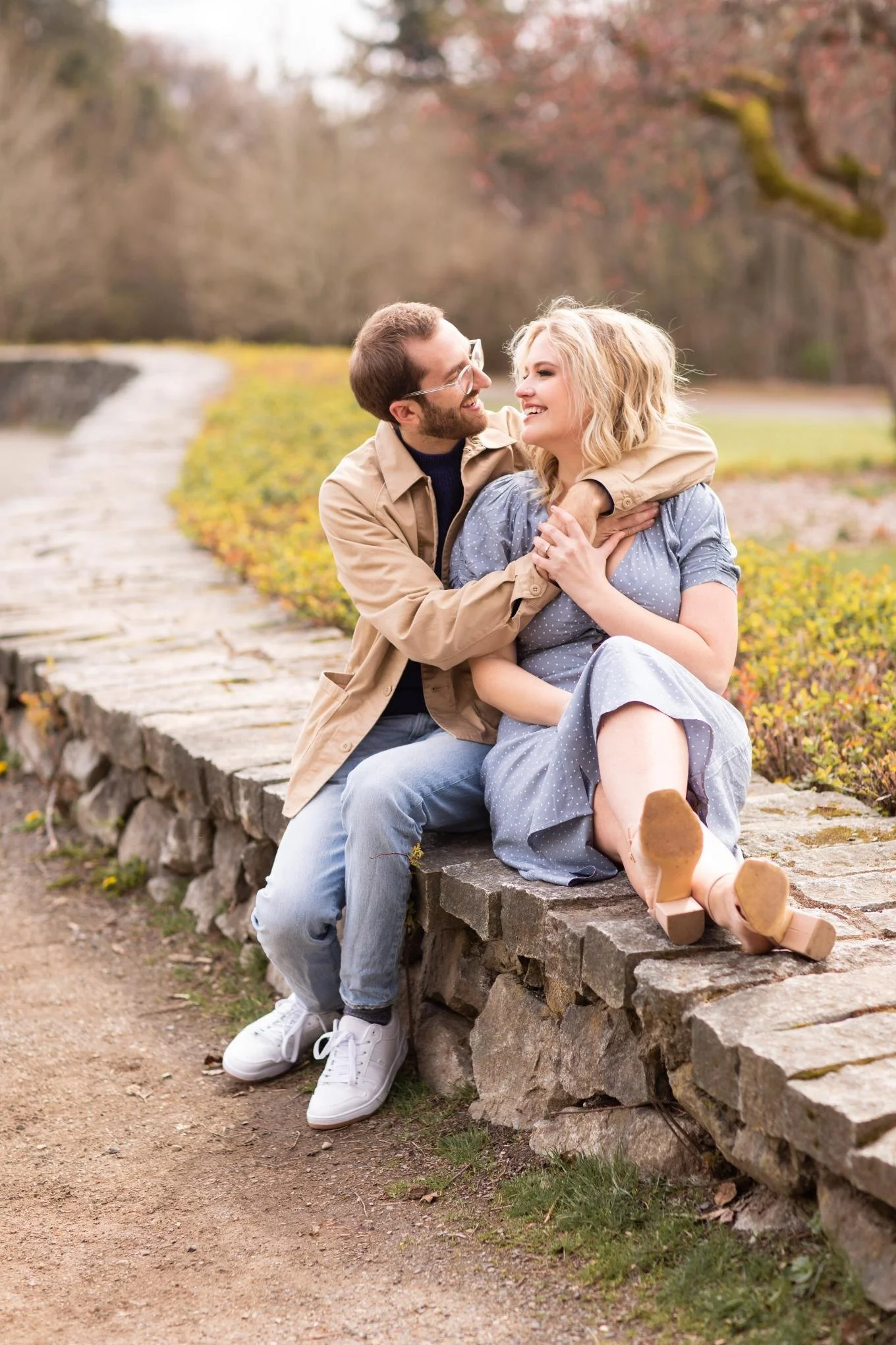 A couple sitting on a stone ledge outdoors during fall, smiling, with the man playfully wrapping his arms around the woman, who is barefoot and wearing a blue polka-dot dress, with leafless trees and a winding path in the background.