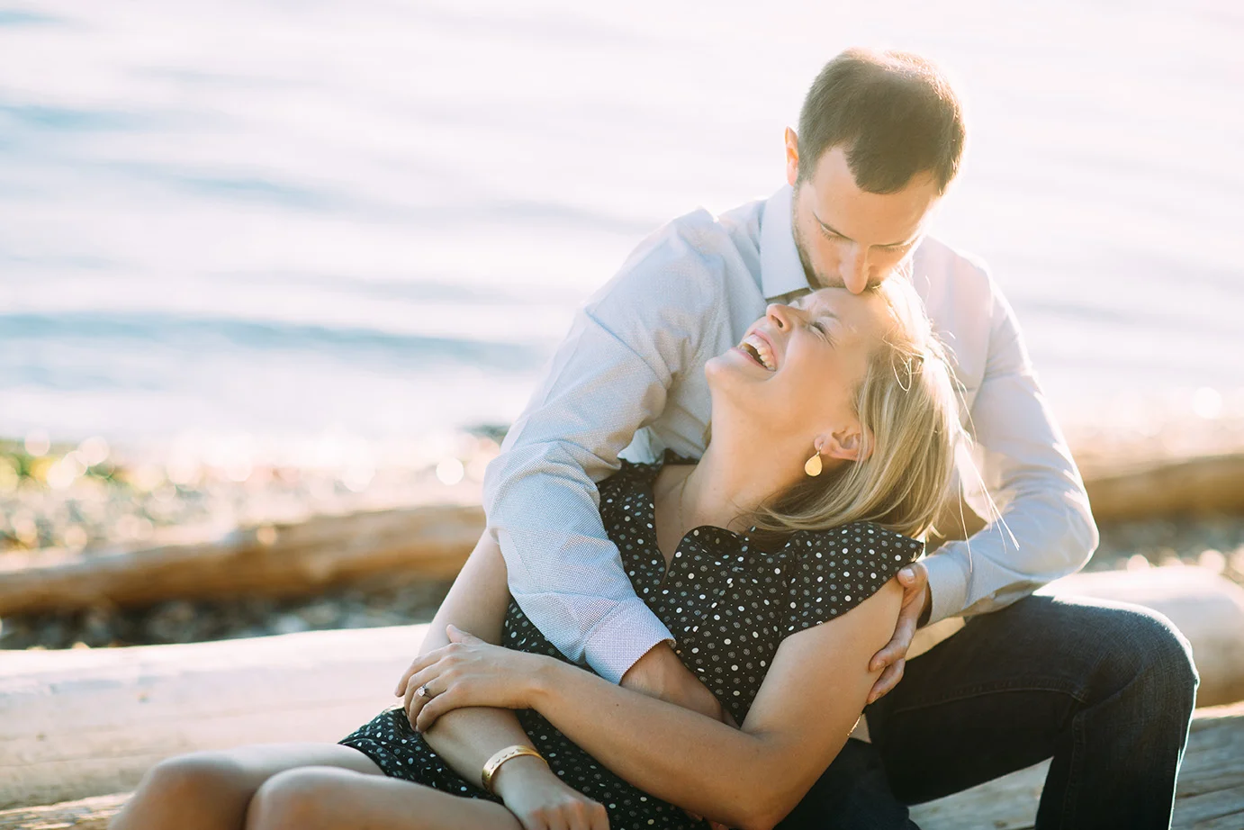 A couple enjoying a joyful moment on the beach in West Seattle at Lincoln Park, with the man kissing the woman's forehead as they sit on a log near the water.