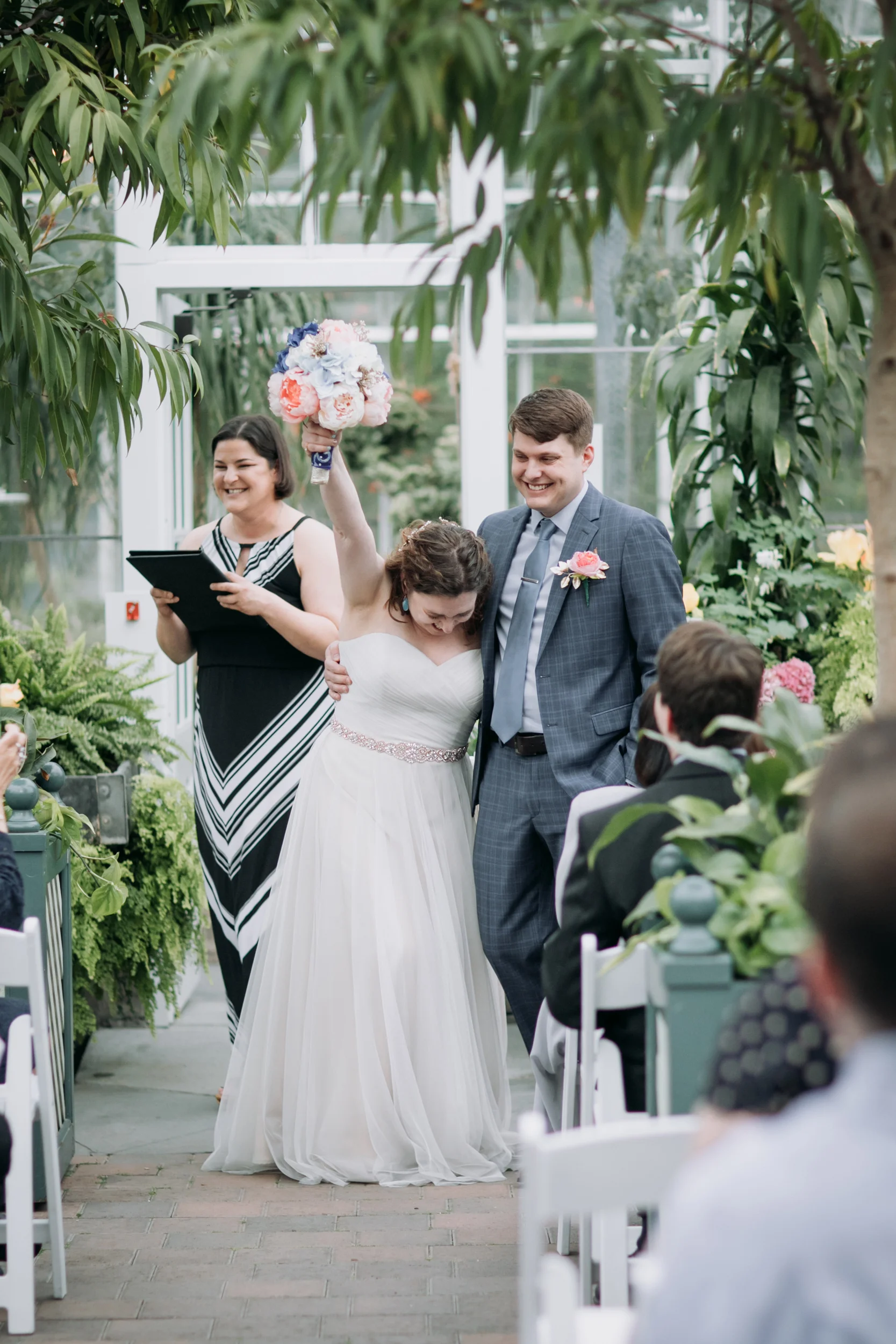 A bride and groom celebrating during their wedding ceremony inside a greenhouse, with the bride holding a colorful bouquet, and guests seated in front of them.