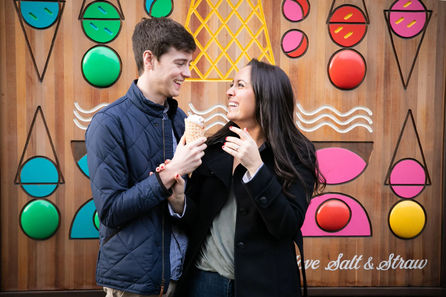 A young couple sharing an ice cream cone in front of a colorful, geometric wall art with circles, lines, and shapes. The man is wearing a navy quilted jacket and smiling, while the woman is wearing a black coat and playfully touching her hair.
