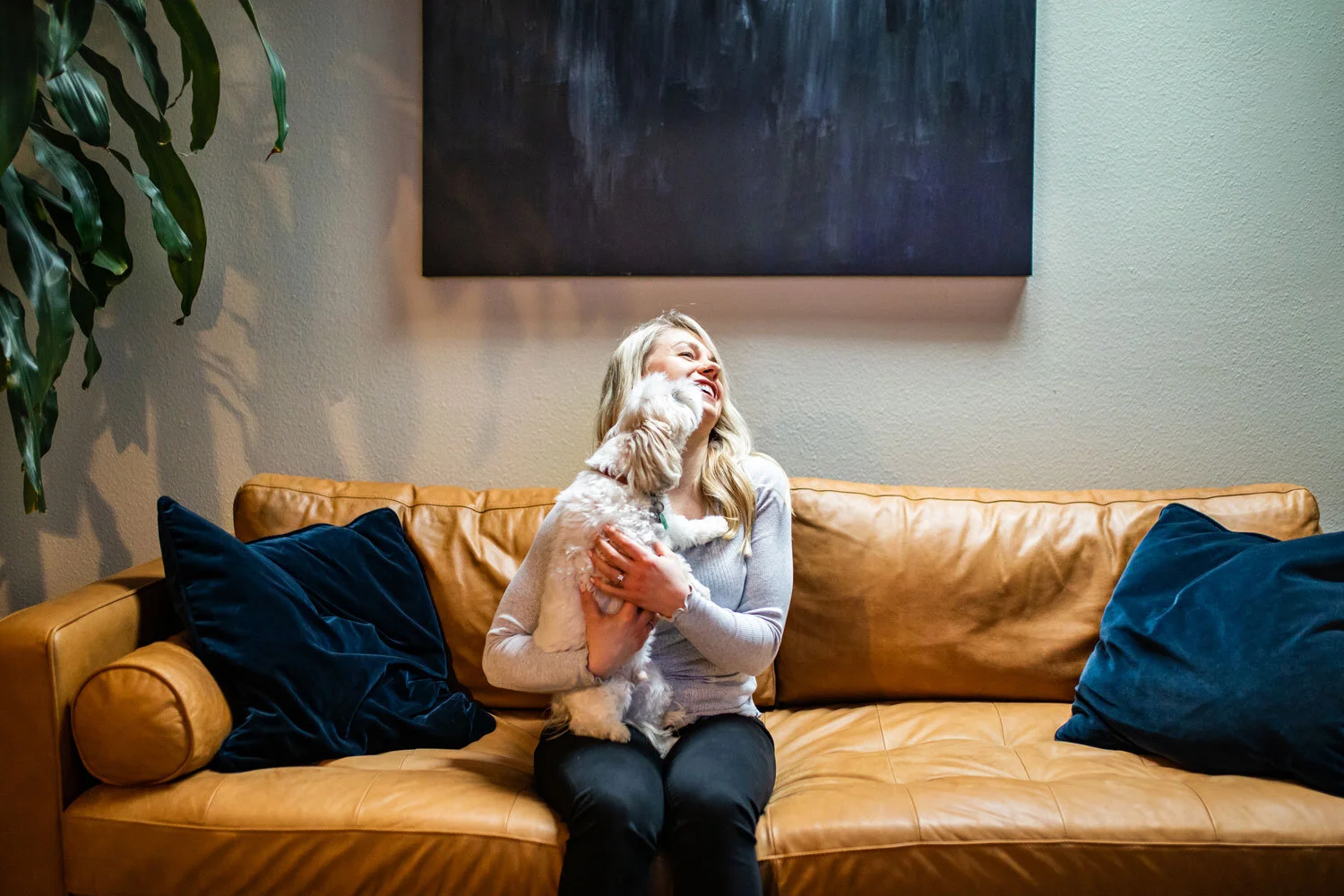 A woman with blonde hair sitting on a tan leather couch, holding a small fluffy dog and smiling joyfully. The couch has two dark blue pillows, and there is a tall green plant to the left and a dark abstract painting on the wall behind her.