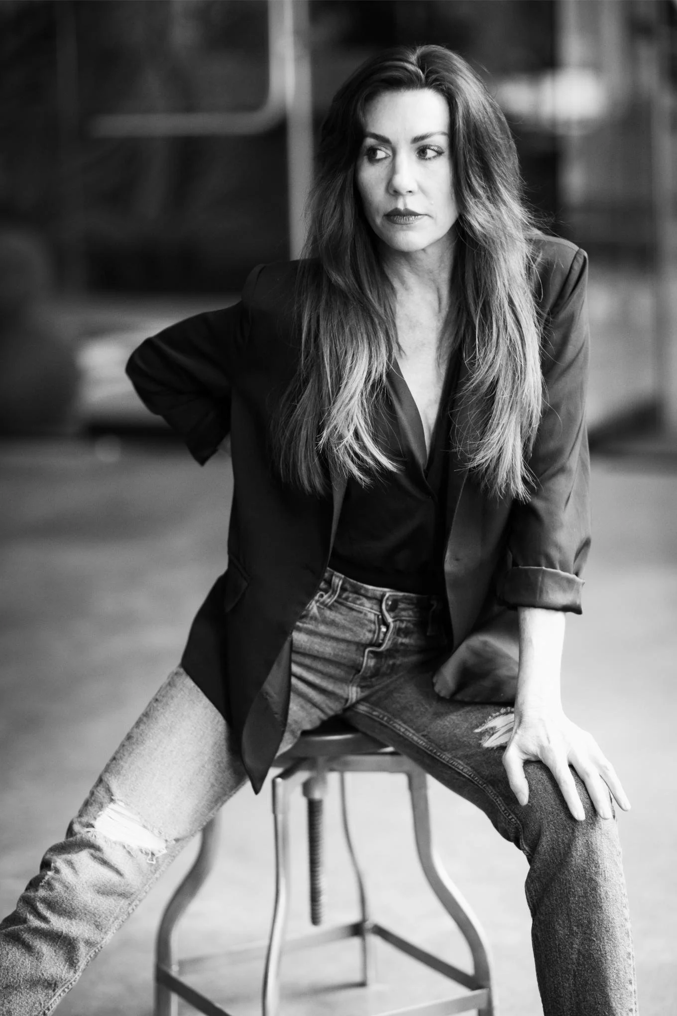 Black and white photo of a woman with long, wavy hair, wearing a blazer and ripped jeans, seated on a stool with one arm resting on her knee, looking to the side with a serious expression.