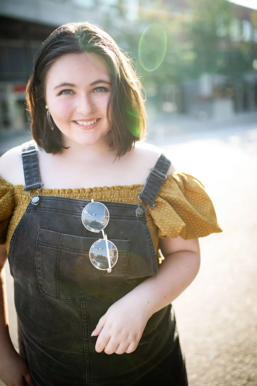 Smiling young woman with shoulder-length dark hair wearing a mustard yellow top with puffed sleeves and black overalls, standing outdoors in bright sunlight with sunglasses hanging on her overalls.