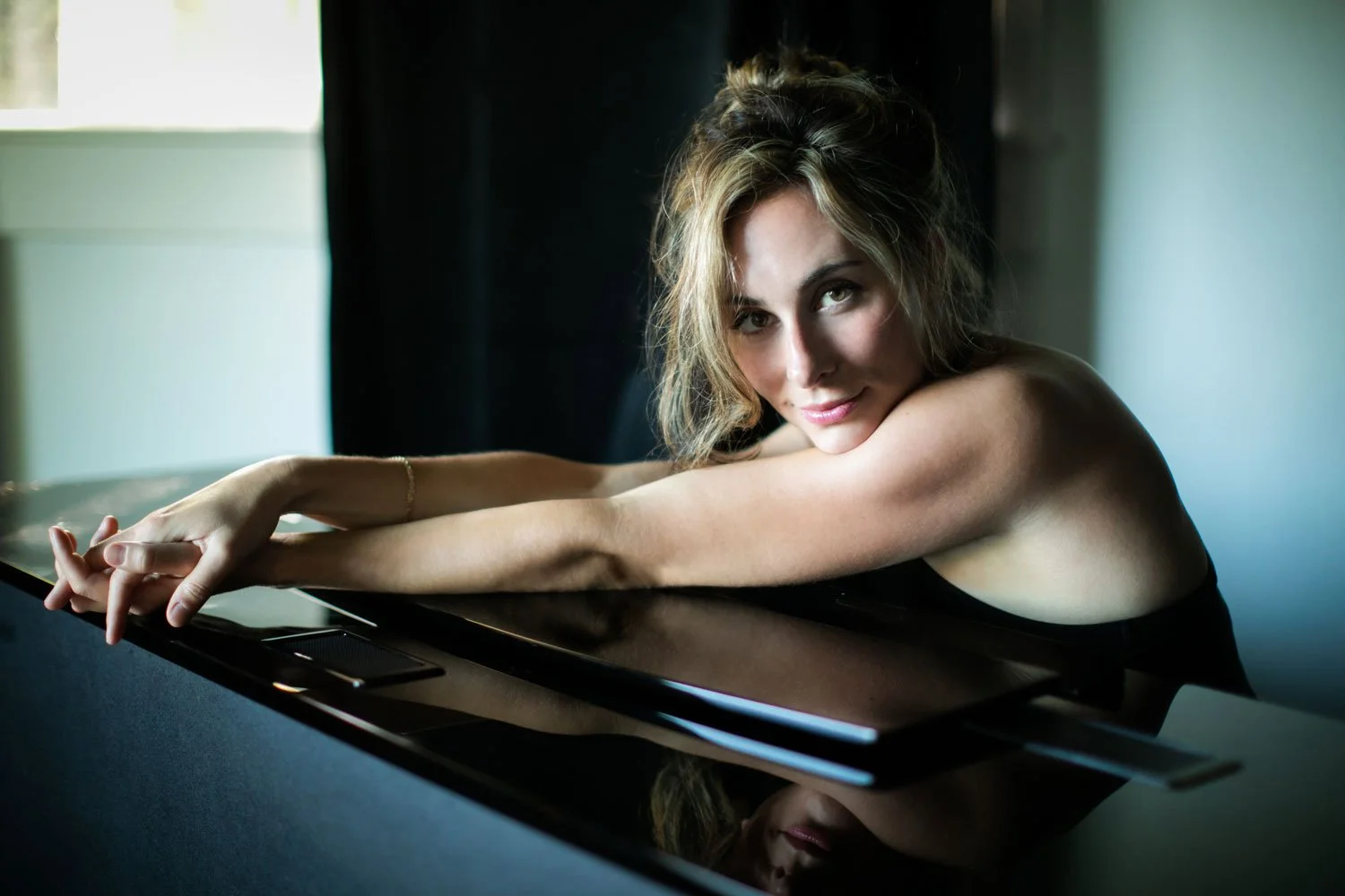 A woman with wavy hair resting her arms on a reflective black surface, looking at the camera with a slight smile in a softly lit room.