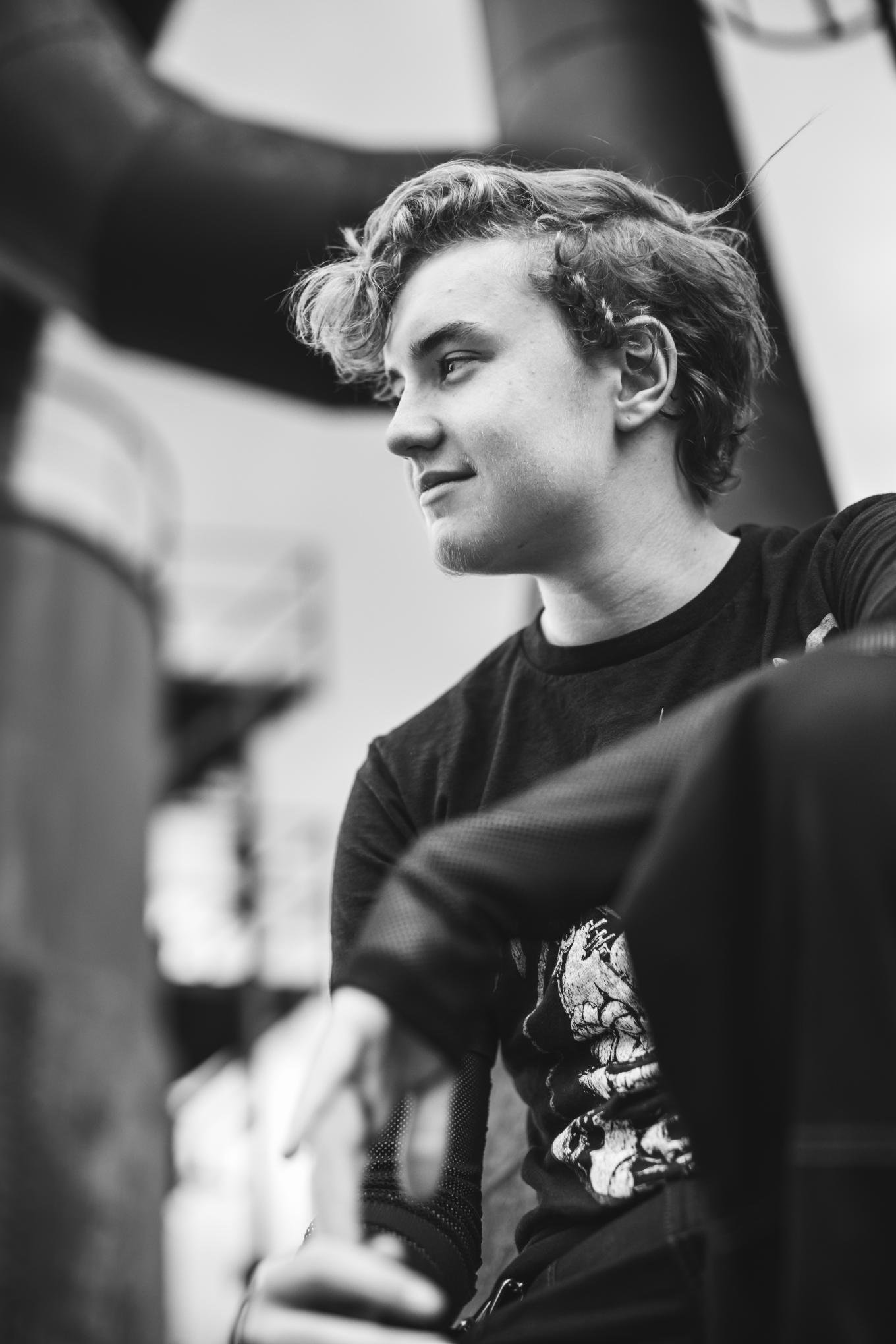 A young man with curly hair and a faint beard, wearing a graphic T-shirt, is sitting and looking down with a slight smile. The photo is in black and white, and there is a blurred person in the foreground wearing glasses and a jacket.