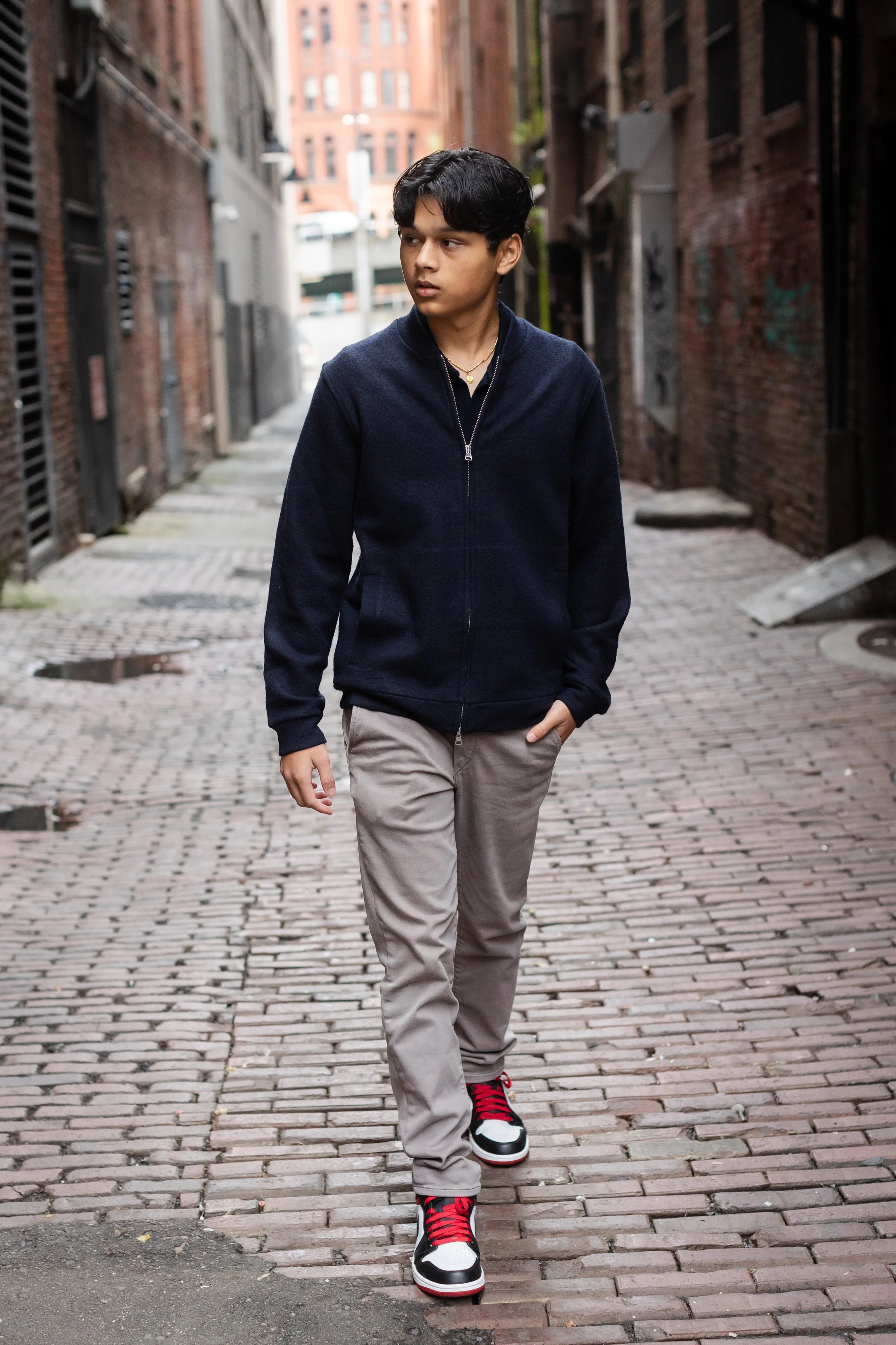 A young man with dark hair walking down a cobblestone alleyway, wearing a navy blue zip-up jacket, beige pants, and red and black sneakers.