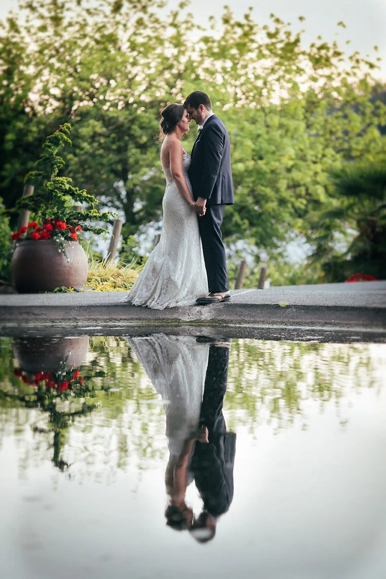 A bride and groom stand close together by a winery pond in Seattle, their reflection visible in the water, with lush green trees and foliage in the background.