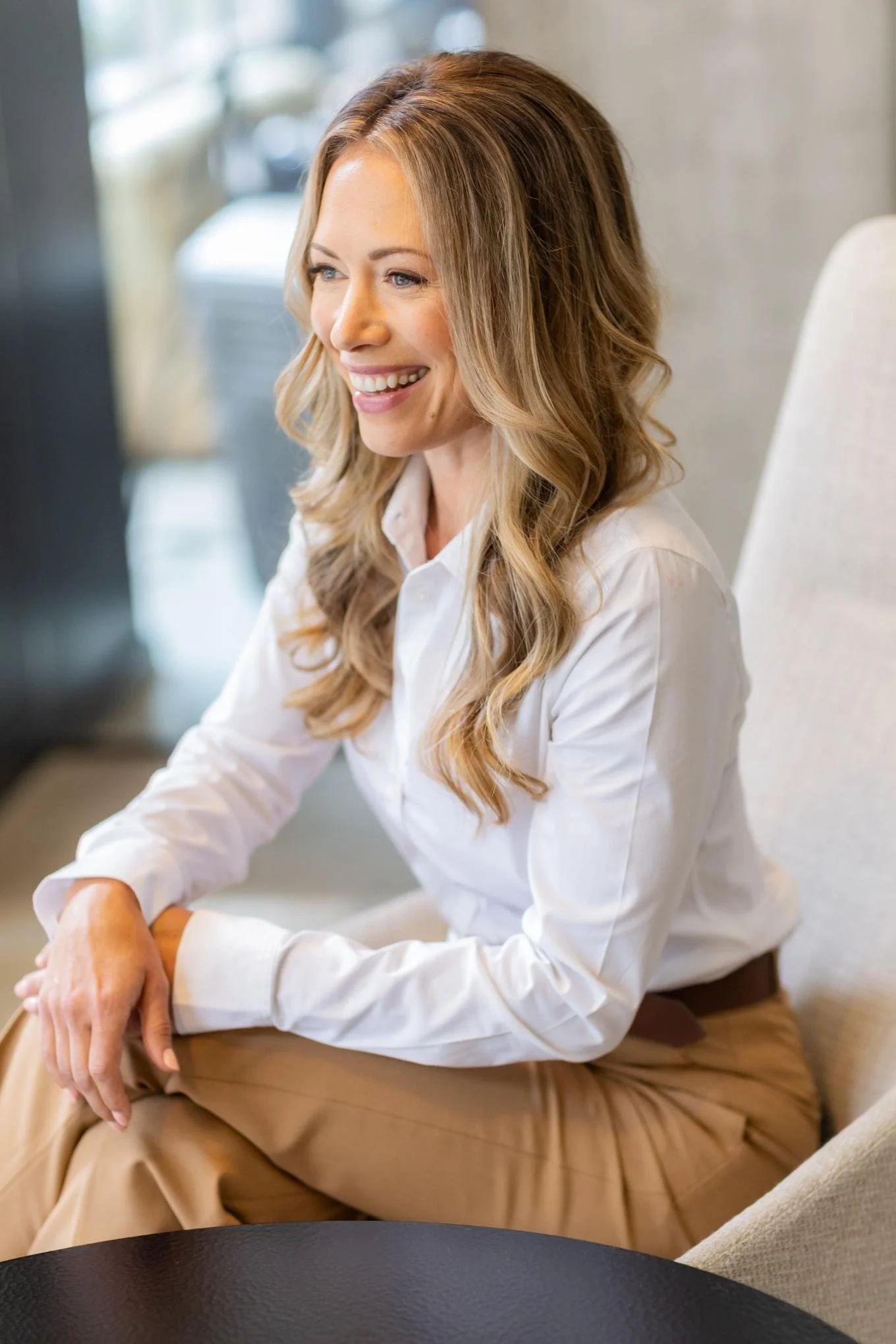 A woman with long wavy blonde hair sitting on a beige chair, smiling, wearing a white blouse and beige pants, in a well-lit indoor setting.
