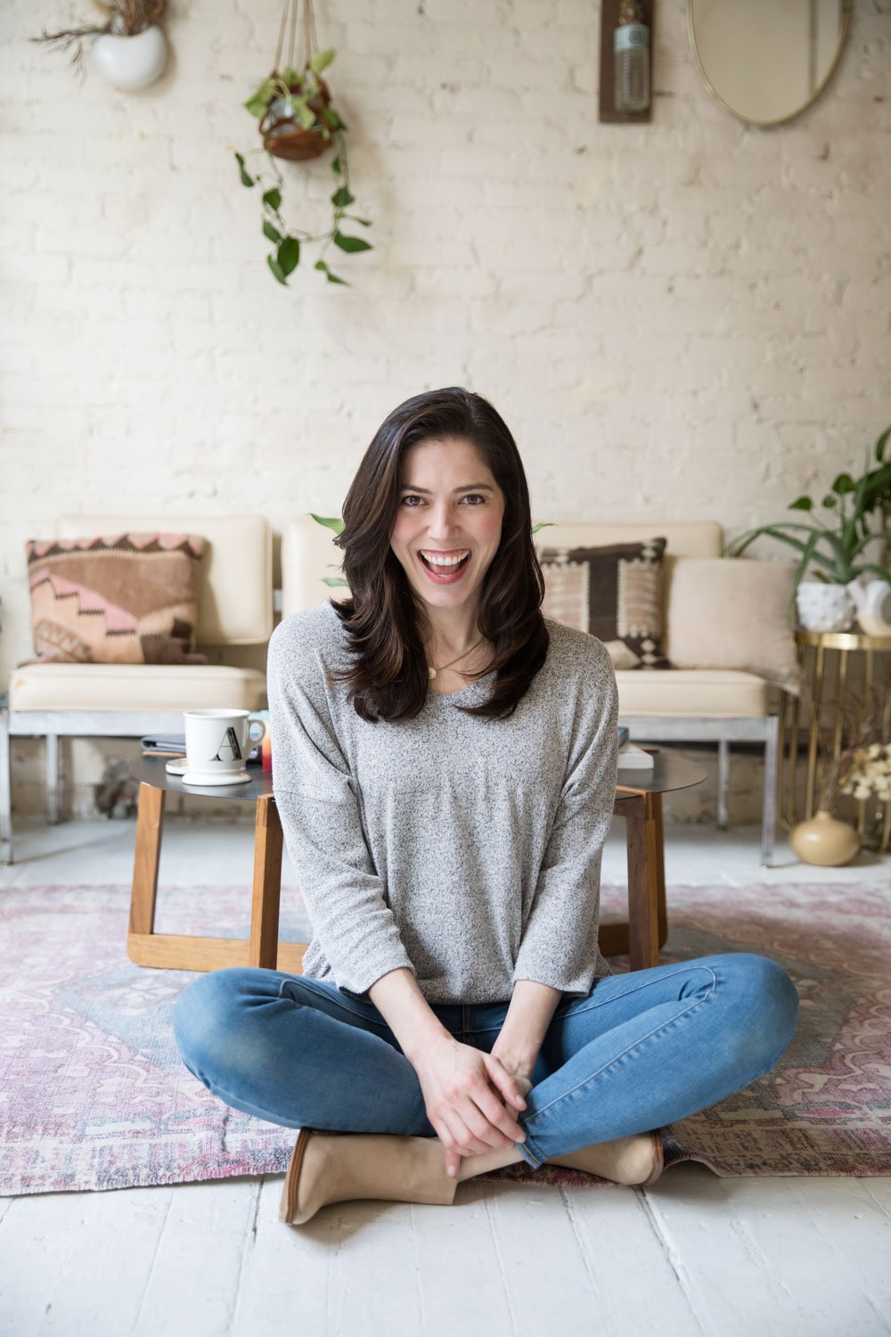 A woman sitting cross-legged on a wooden floor in a cozy living room, smiling and looking at the camera. She has dark hair, is wearing a grey long-sleeved top and blue jeans, with a white mug and a smartphone on a low table behind her. The background