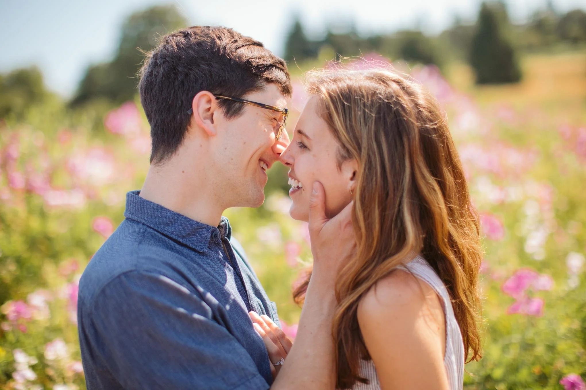 A couple with light skin is smiling and touching noses in a garden with pink flowers, bright sunlight, and greenery in the background.