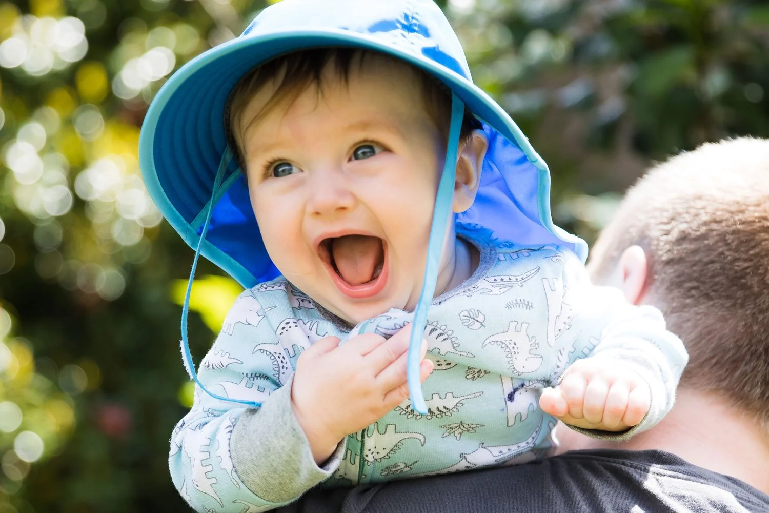 A joyful baby boy with blue eyes and light brown hair laughing while being carried on an adult's shoulder, wearing a blue hat and a dinosaur-patterned shirt, outdoors with blurred greenery in the background.