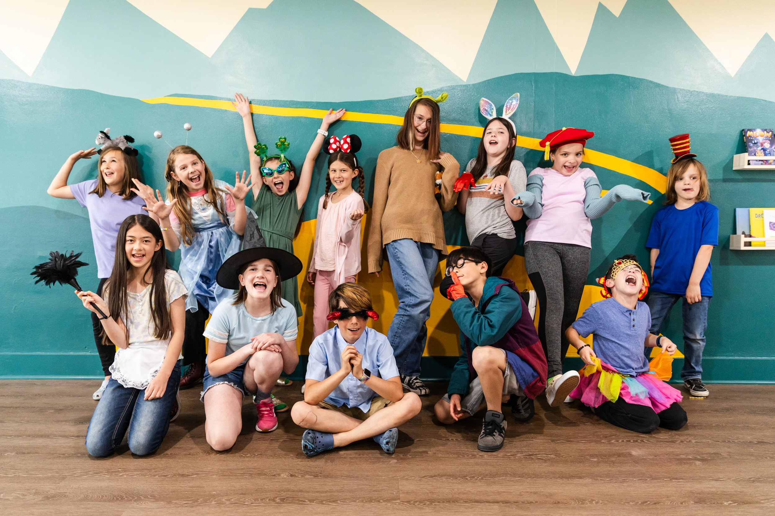 Children in colorful costumes and accessories posing and laughing in front of a painted mountain mural during a fun event.
